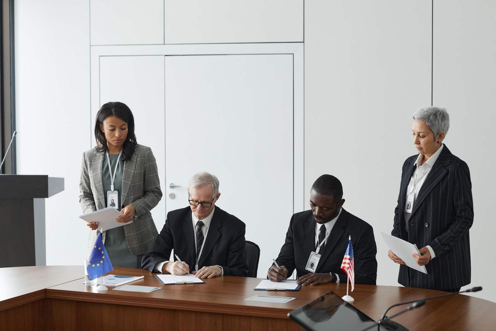 Two officials sign documents at a table bearing European Union and American flags while colleagues review papers standing behind them
