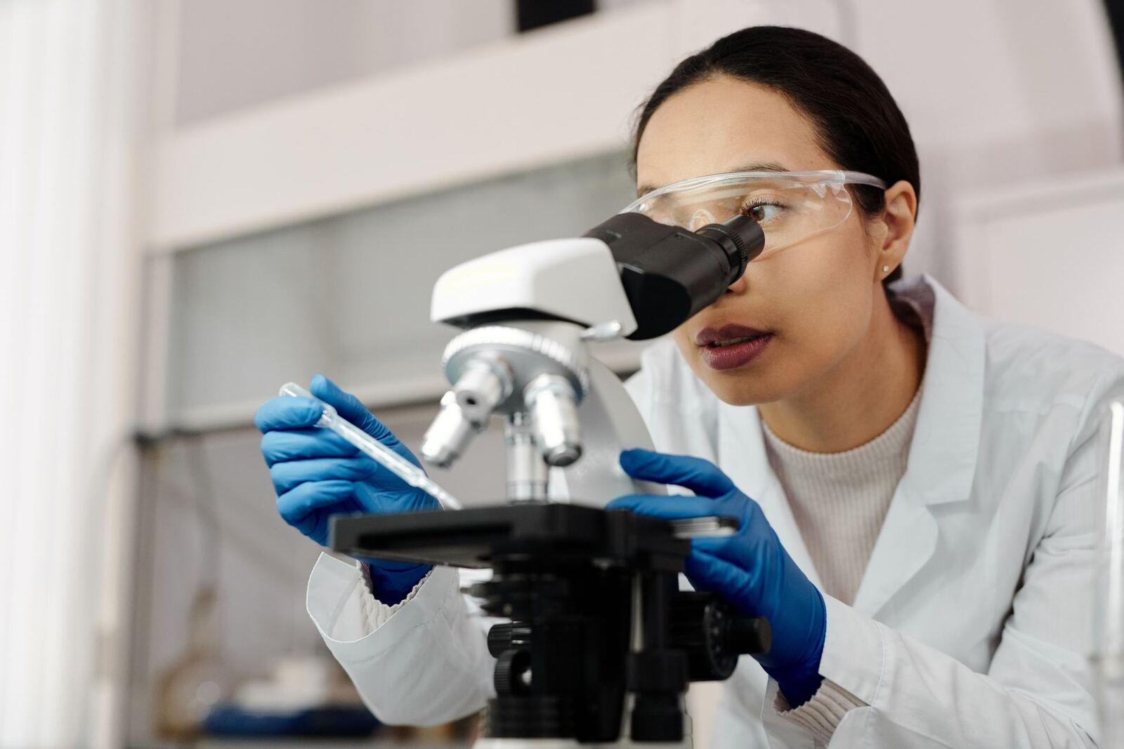 A scientist in a lab coat and safety glasses examines a sample through a microscope while holding a pipette