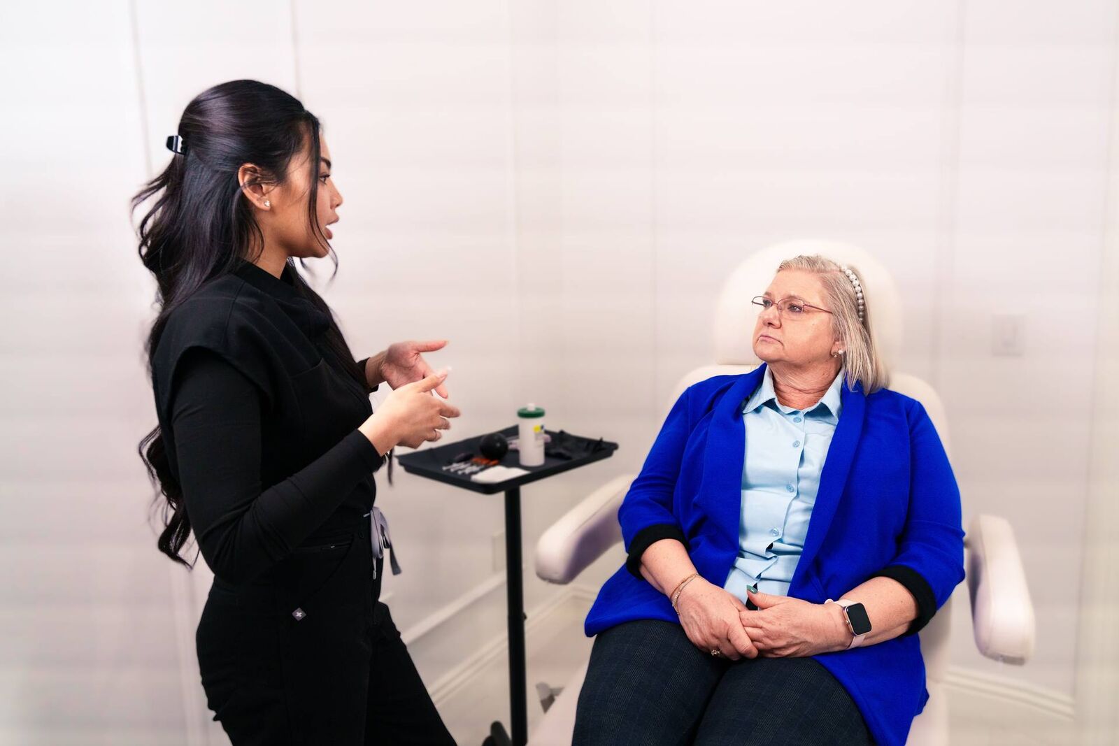 A clinician in a dark top speaks with a seated patient during a consultation in a bright exam room
