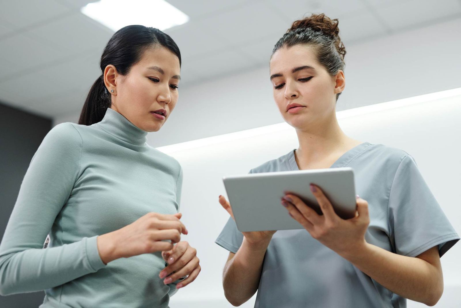A nurse holding a tablet consults with a colleague in a bright white clinic hallway
