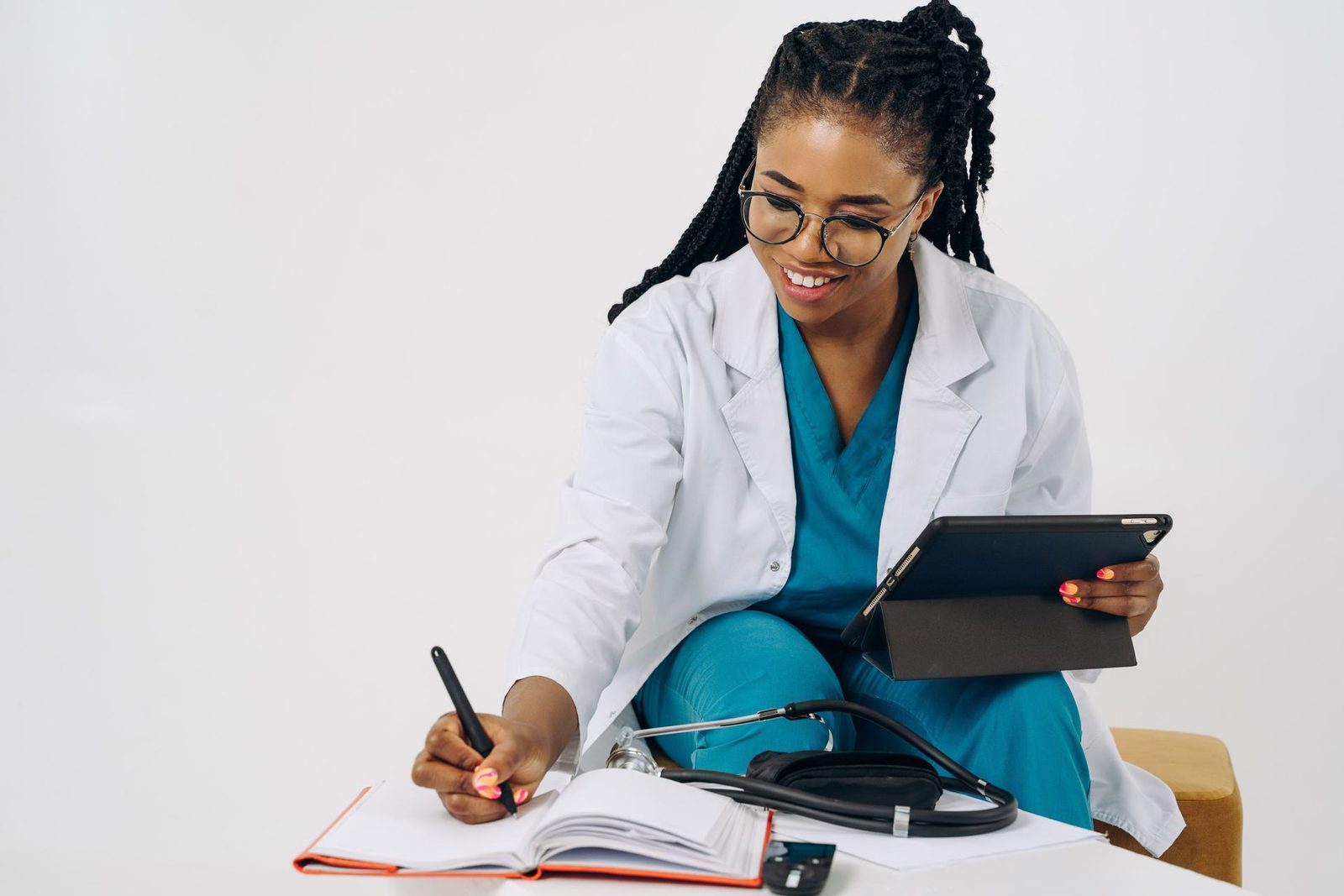 A physician in a white coat and scrubs writes in a notebook beside a stethoscope and a tablet