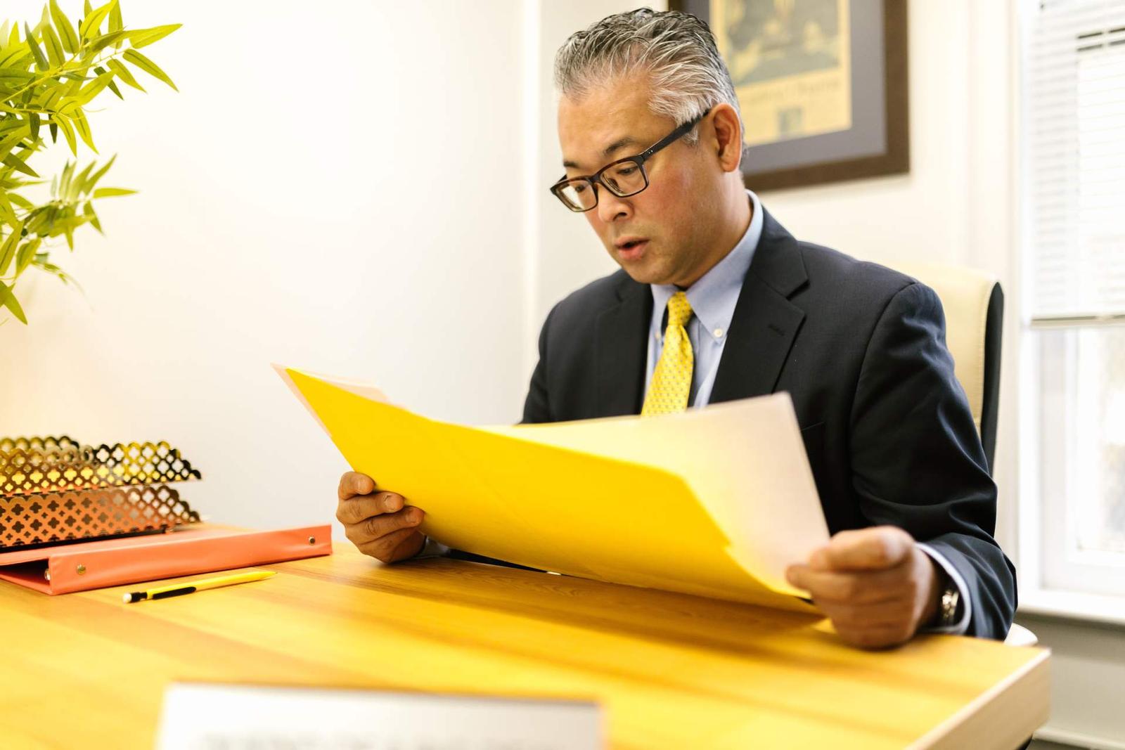 A man in a dark suit reads intently from an open yellow folder at a wooden desk