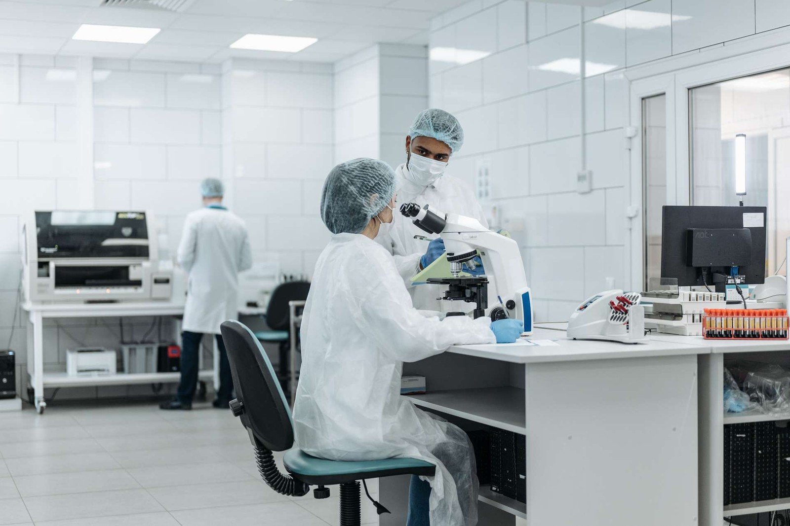 Two lab technicians in cleanroom garb examining a sample under a microscope in a bright diagnostics laboratory