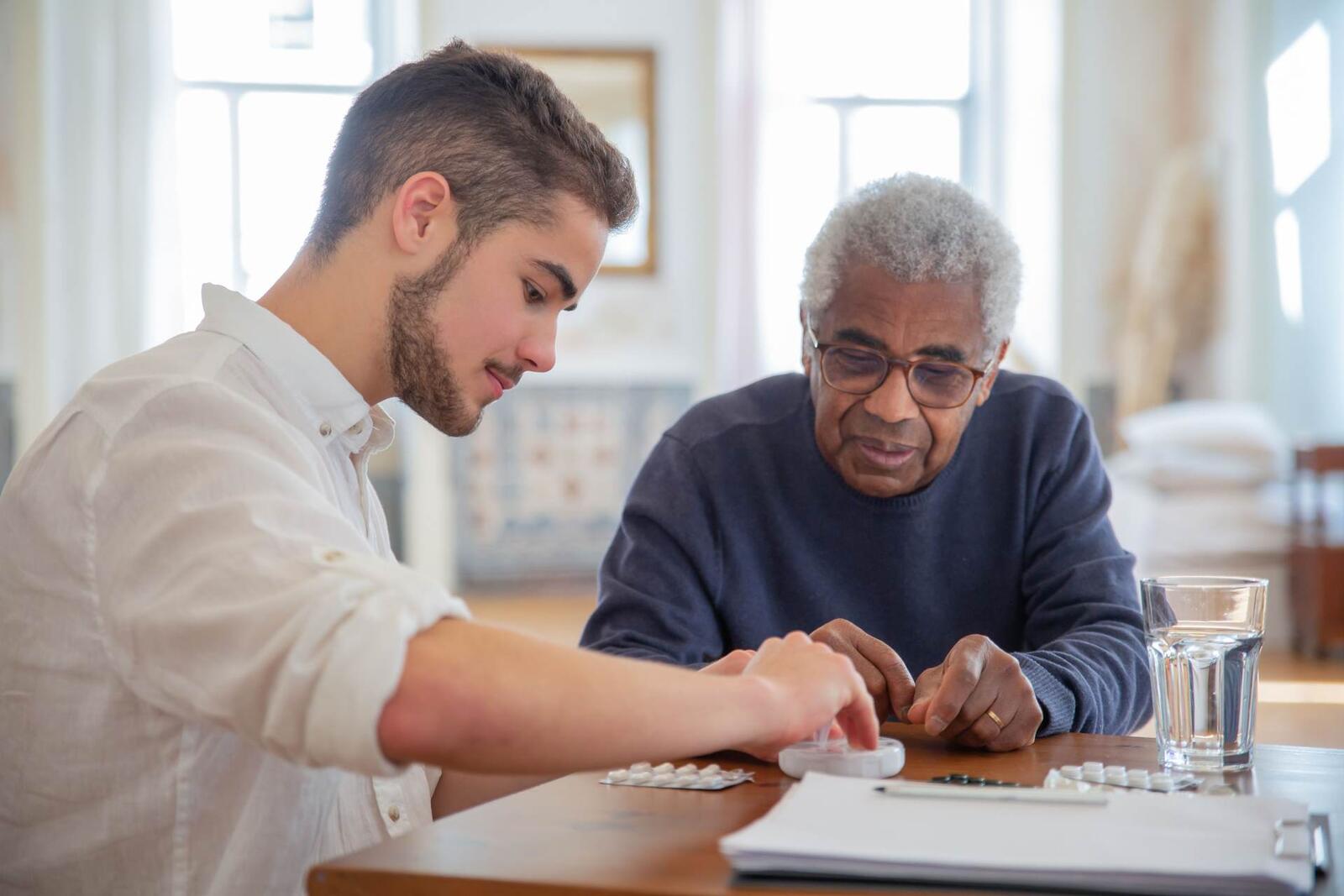 A younger man helps an older man sort prescription medication into a daily pill organizer at a table