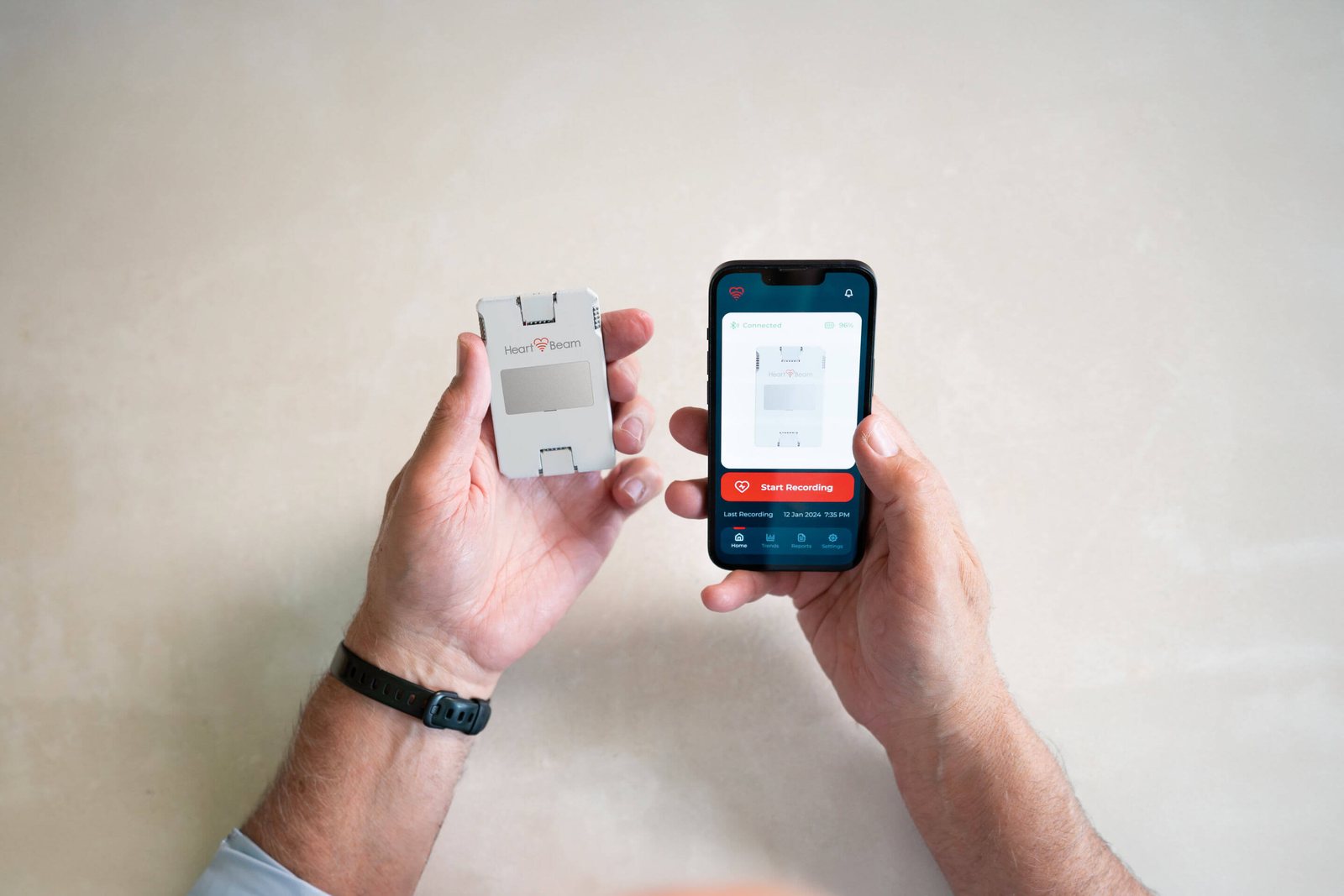 A person holds the credit-card-sized HeartBeam ECG device in one hand and a smartphone running the HeartBeam app, its Start Recording button showing, in the other, over a pale stone surface