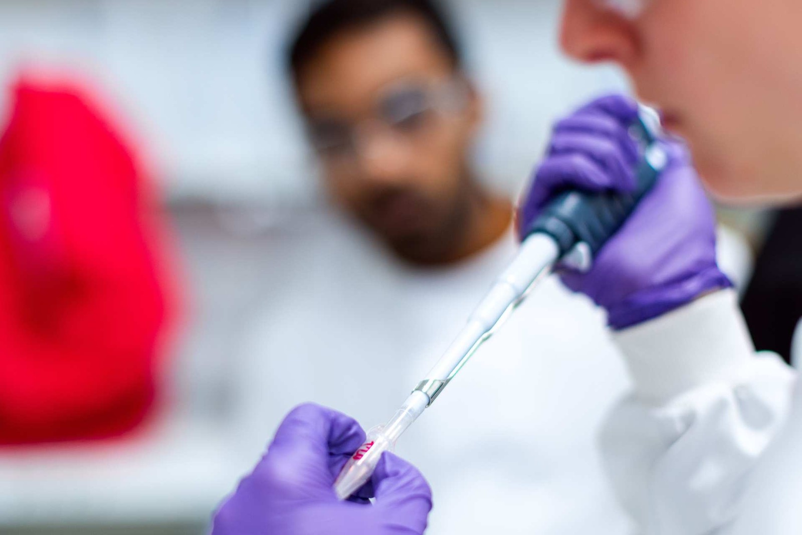 A gloved researcher holds a pipette over a sample tube while a colleague looks on in a laboratory