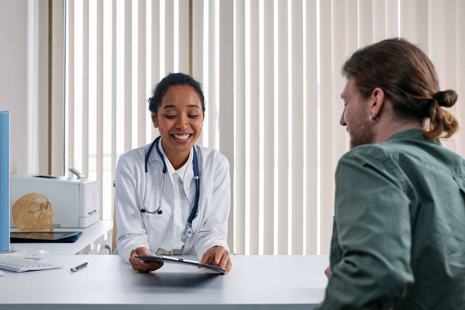 A physician in a white coat and stethoscope reviews a tablet with a patient during a clinic consultation