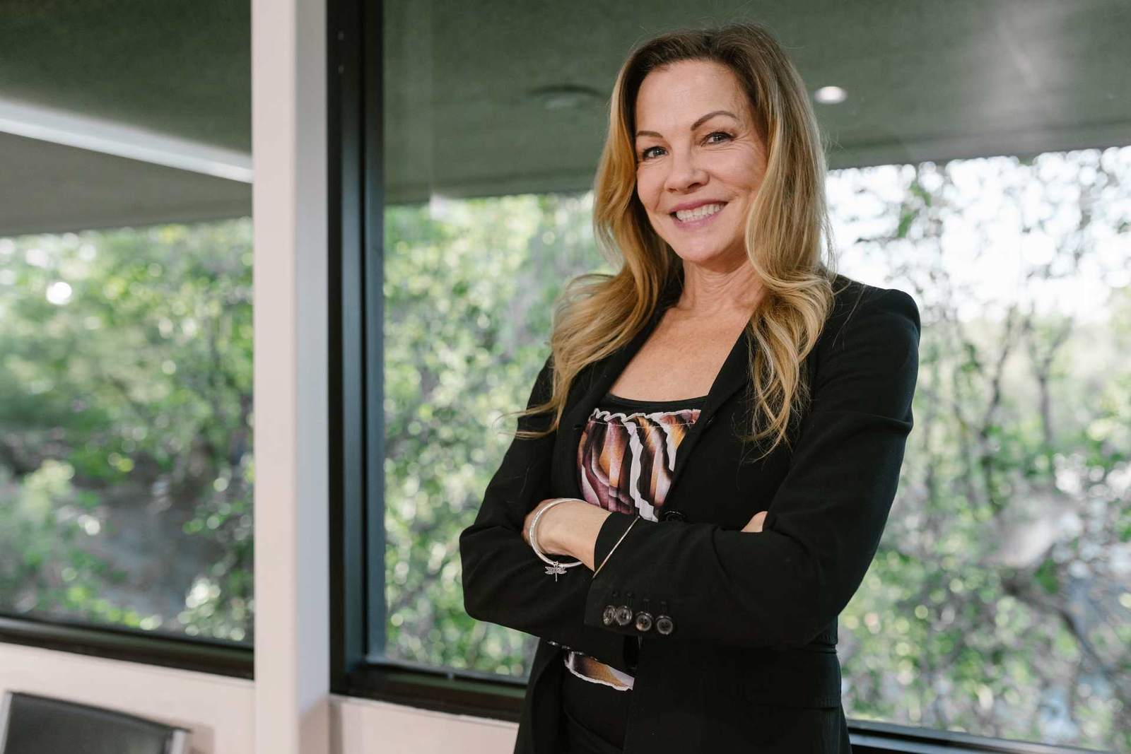 A smiling executive stands with arms crossed in front of a window overlooking trees