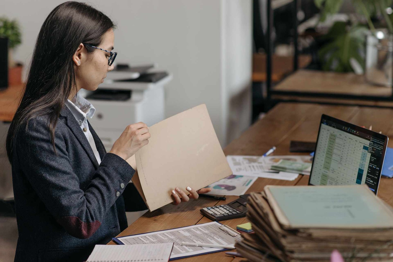 A woman in an office reviews a paper case folder beside a tall stack of aged file records and a laptop