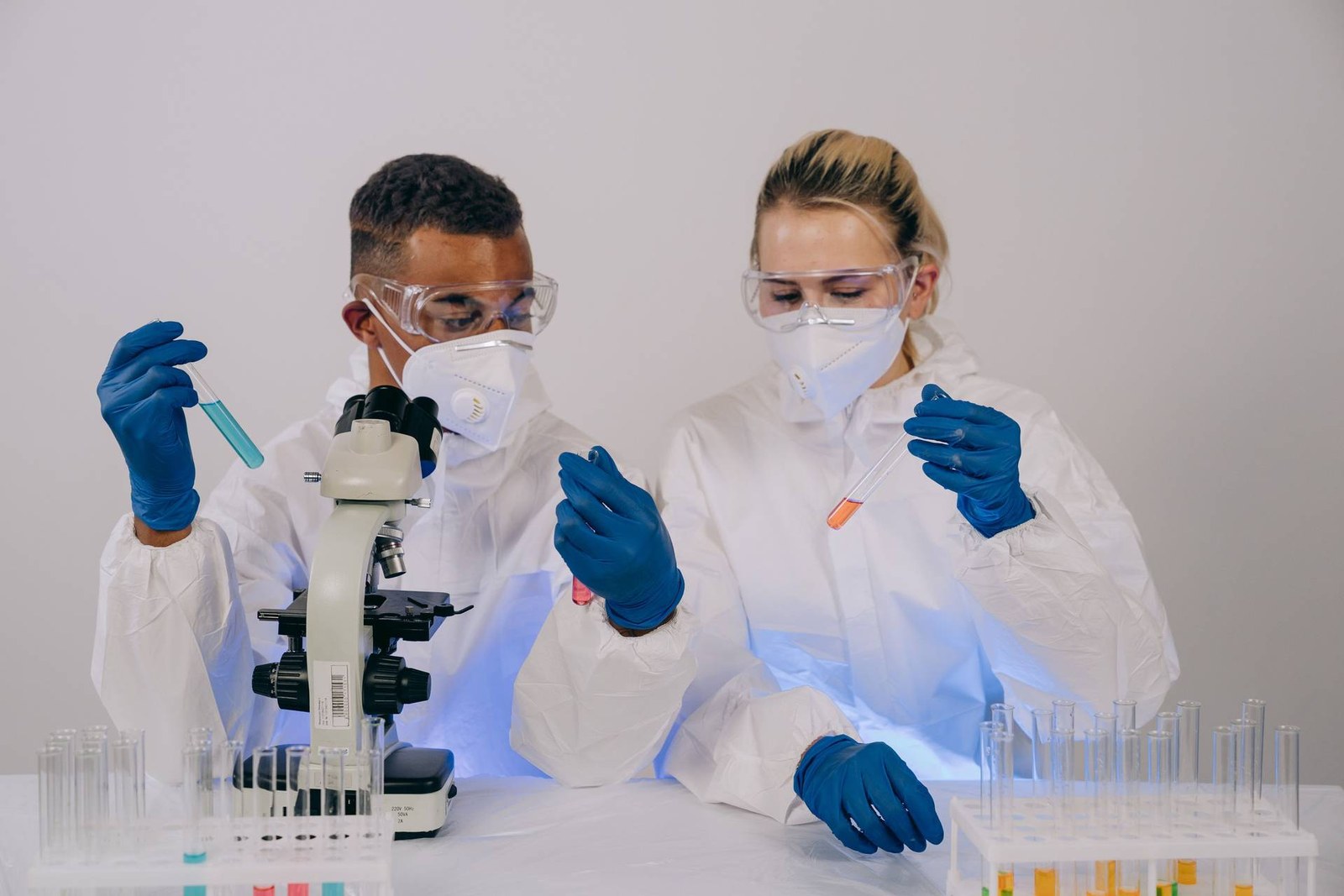 Two masked lab scientists in white coats and safety goggles examine test tubes beside a microscope