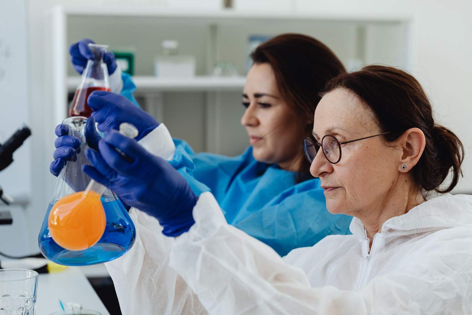 Two scientists in protective gowns and blue gloves hold up an Erlenmeyer flask of blue and orange liquid in a laboratory