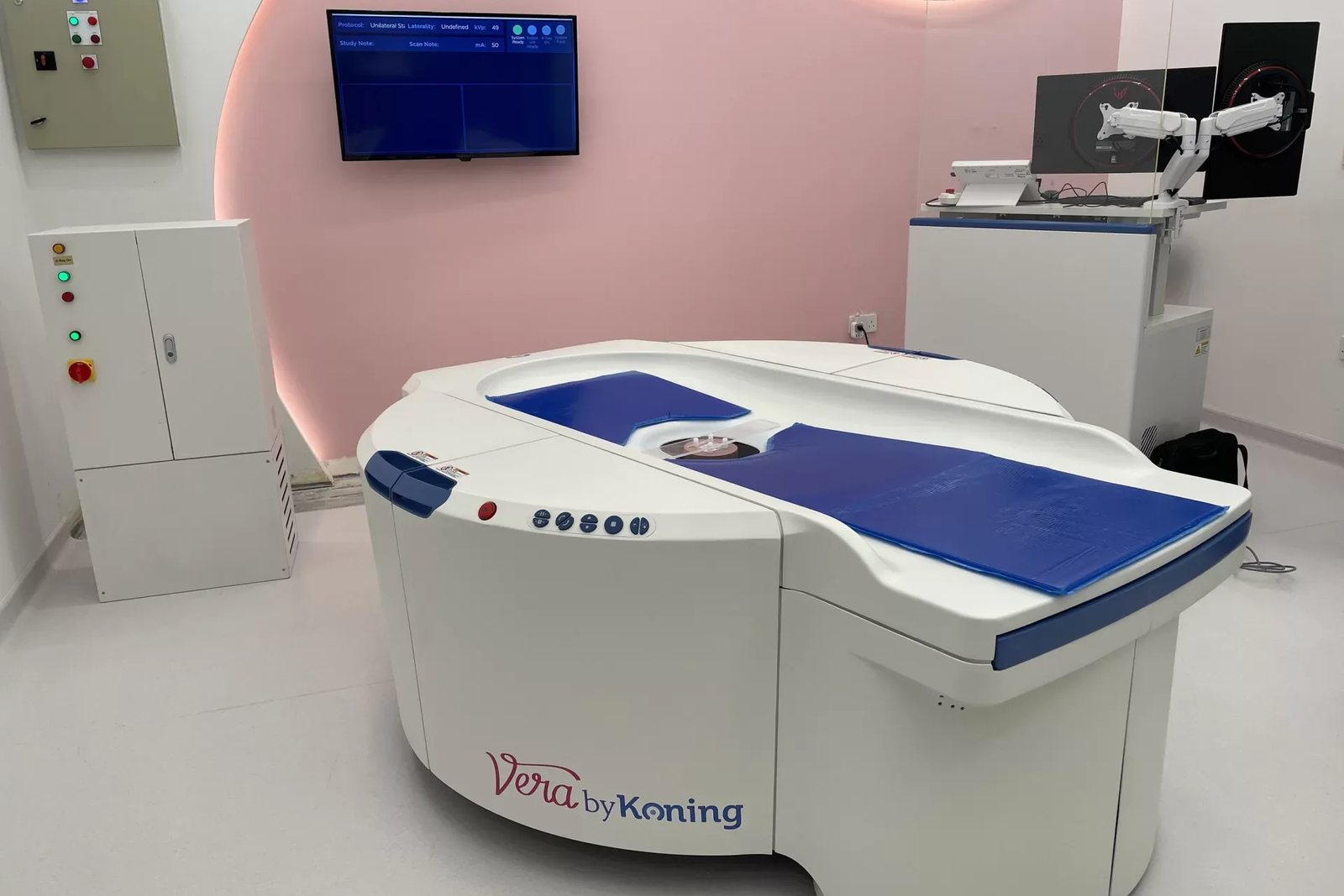 The Koning Vera breast CT scanner installed in an imaging suite: a white, waist-high patient table with a blue cushioned top and a central opening, beside its control cabinet and operator workstation
