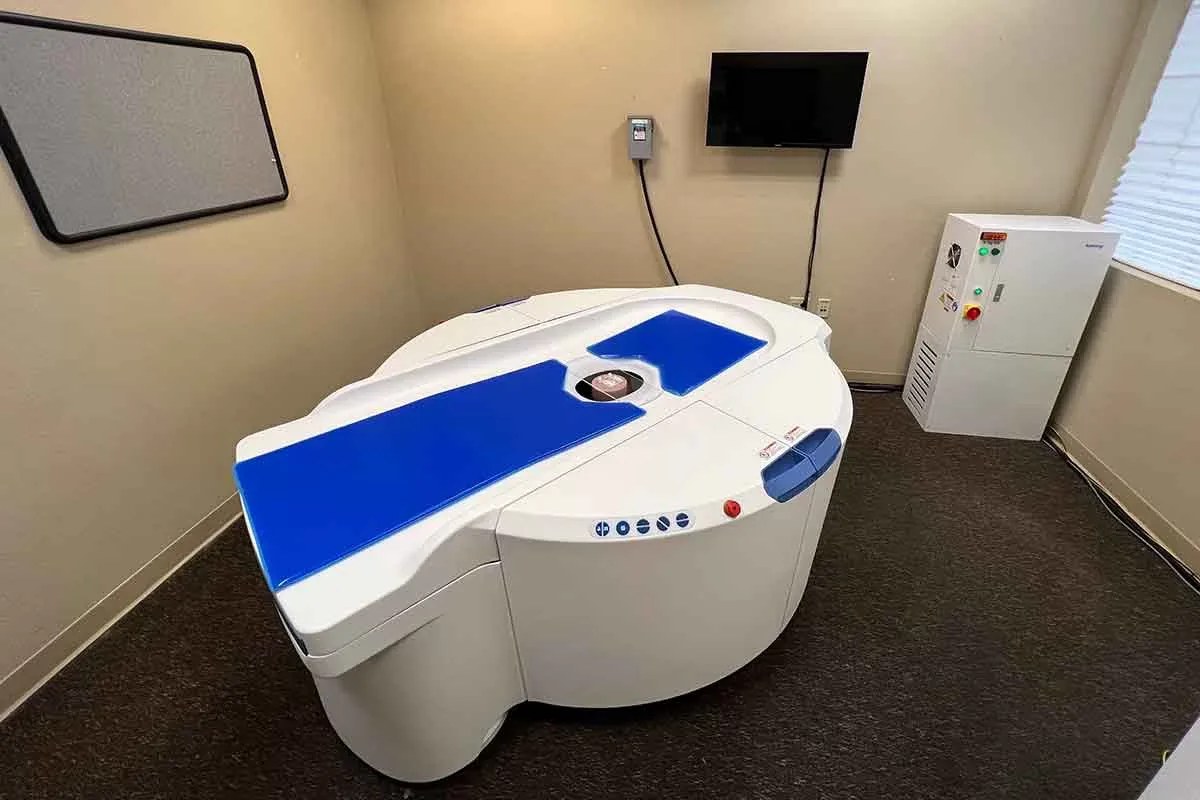 The Koning Breast CT system seen from above in a scan room: the patient lies face down on the blue-padded table with one breast through the central opening, and the scanner rotates beneath