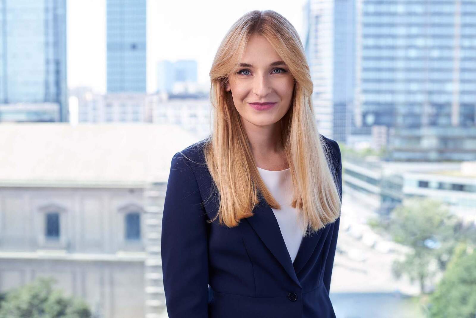 A businesswoman in a navy blazer smiles in an office with a city skyline visible through the window behind her