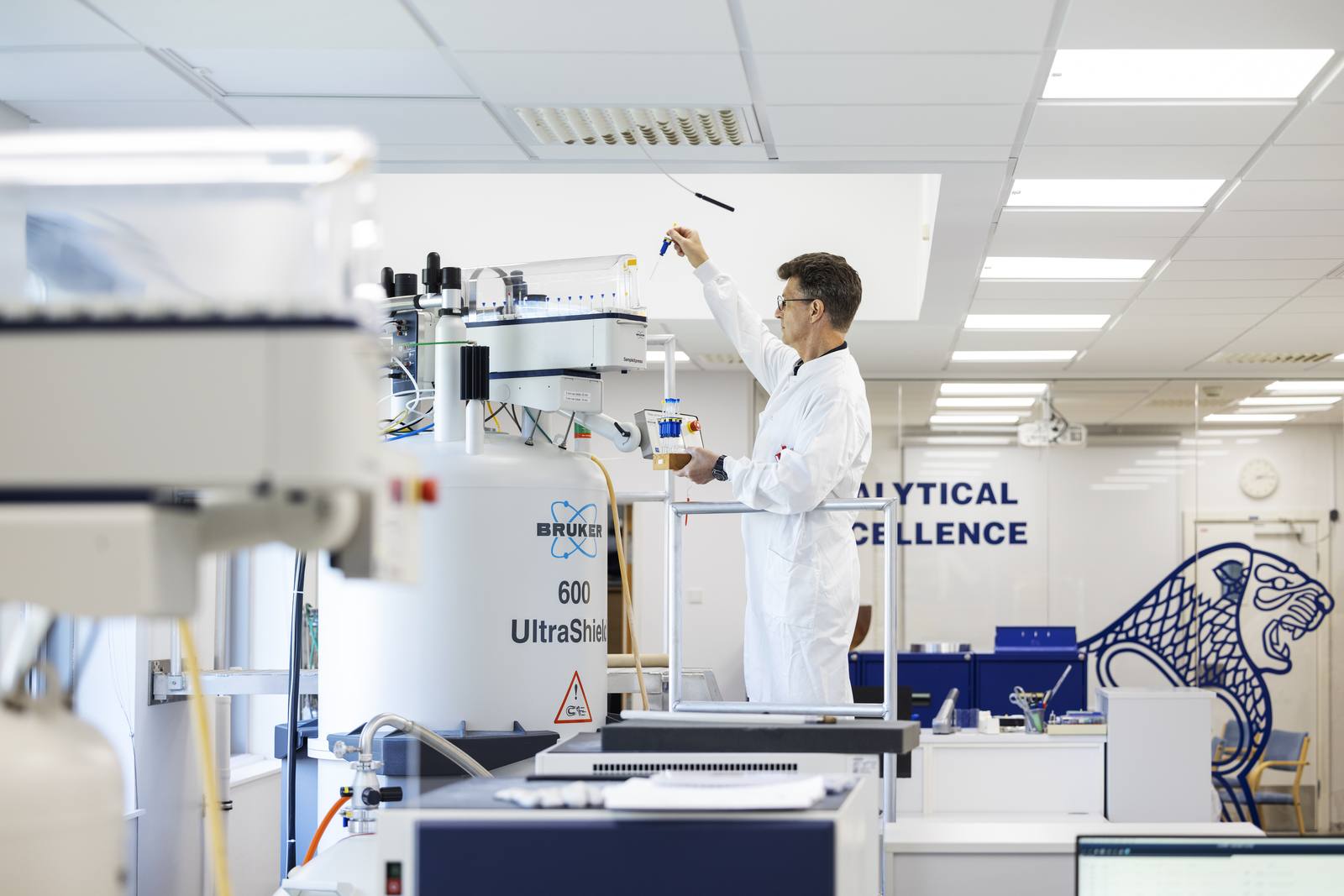 A scientist in a white coat loads a sample into a Bruker 600 UltraShield NMR autosampler in LEO Pharma's analytical laboratory at Ballerup, Denmark, the LEO lion painted on the wall behind.