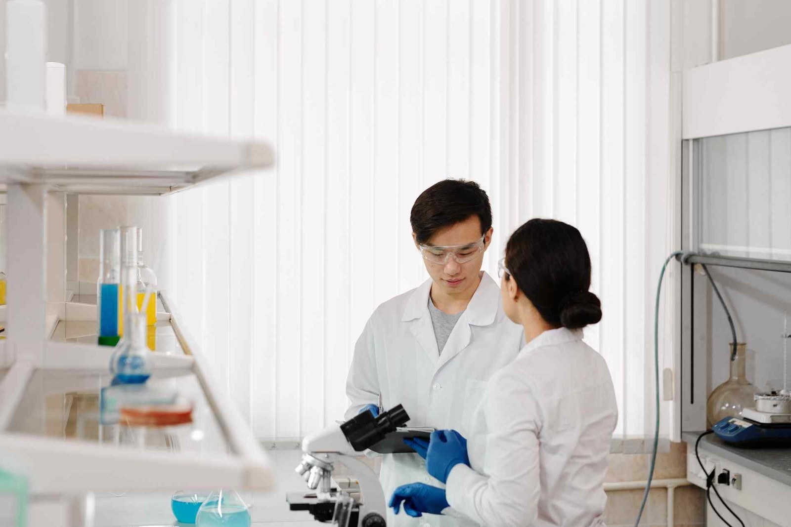 Two researchers in white lab coats and safety goggles review data on a clipboard beside a microscope in a bright laboratory