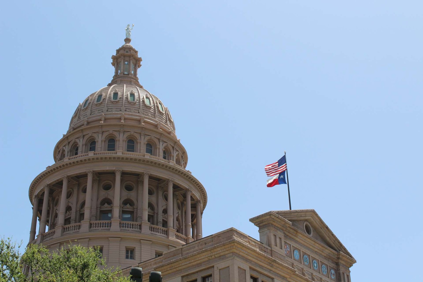 The Texas State Capitol dome against a clear blue sky, with the American and Texas flags flying from an adjacent rooftop