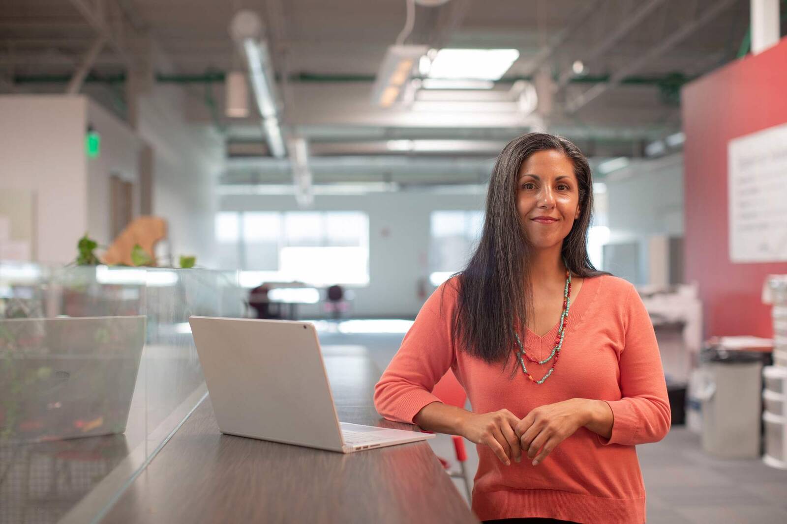 A woman standing at a desk with an open laptop in a bright, loft-style open office
