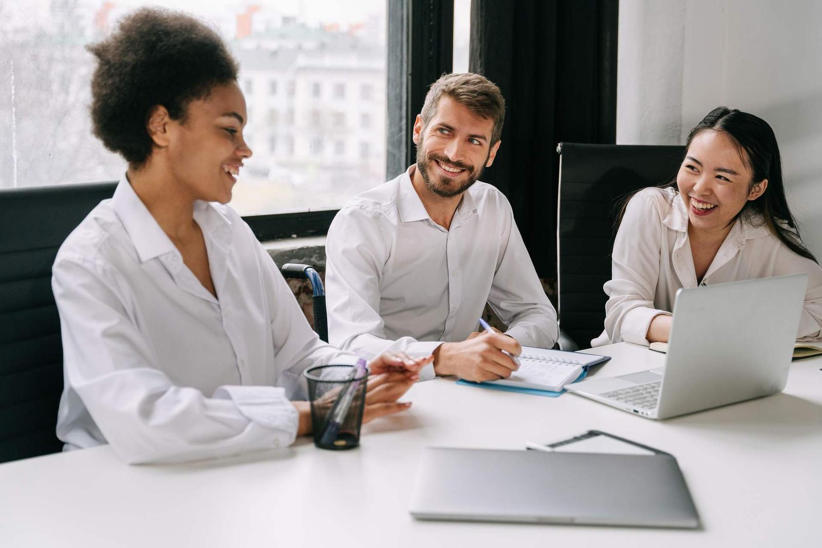 Three colleagues in white shirts share a laugh around a desk with laptops and notebooks open