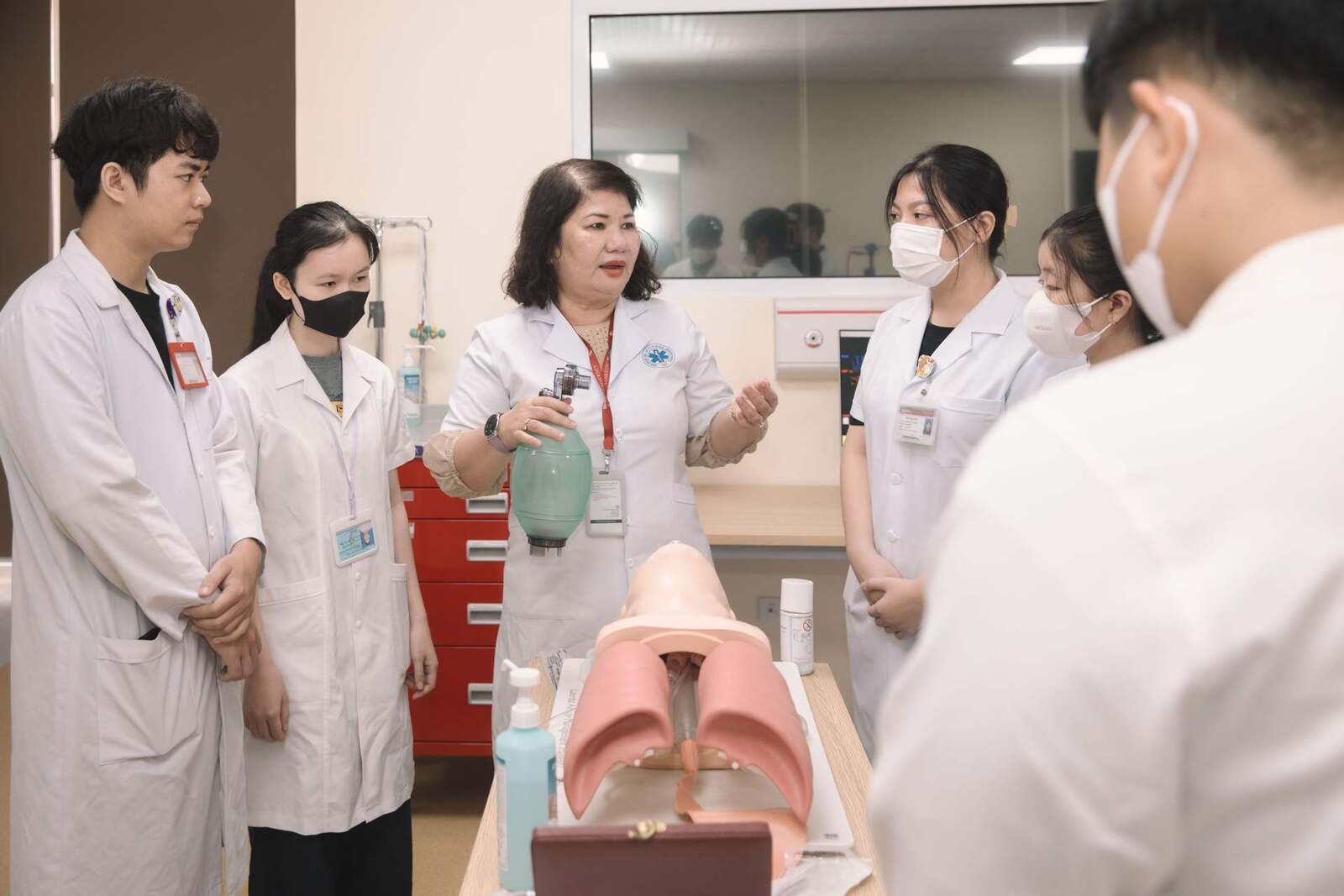 An instructor in a white coat shows a group of masked medical trainees an airway manikin in a clinical training room, a generic stand-in for clinical and medical-education settings