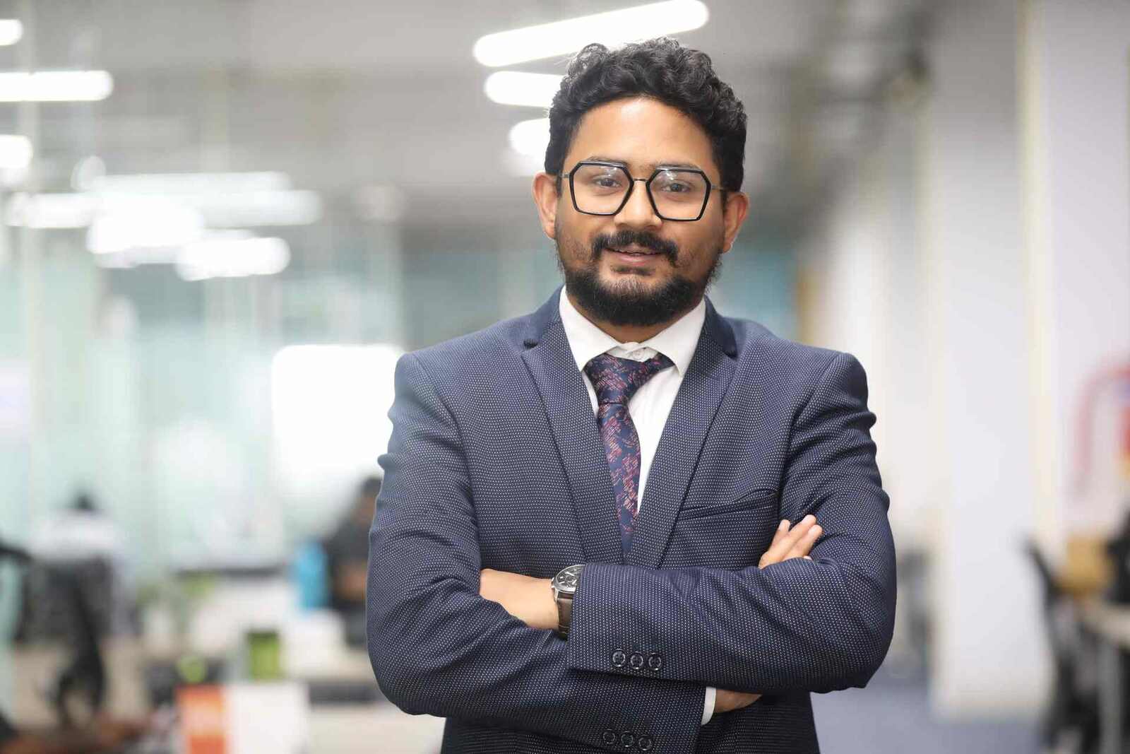 A confident executive in a navy suit, arms crossed, standing in a bright open-plan office