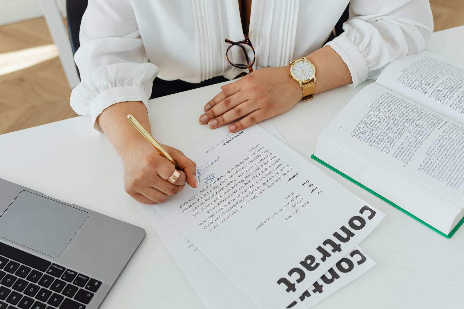 A close-up of a hand in a white blouse signing a contract document with a gold pen, a watch on the wrist, next to a laptop and an open book