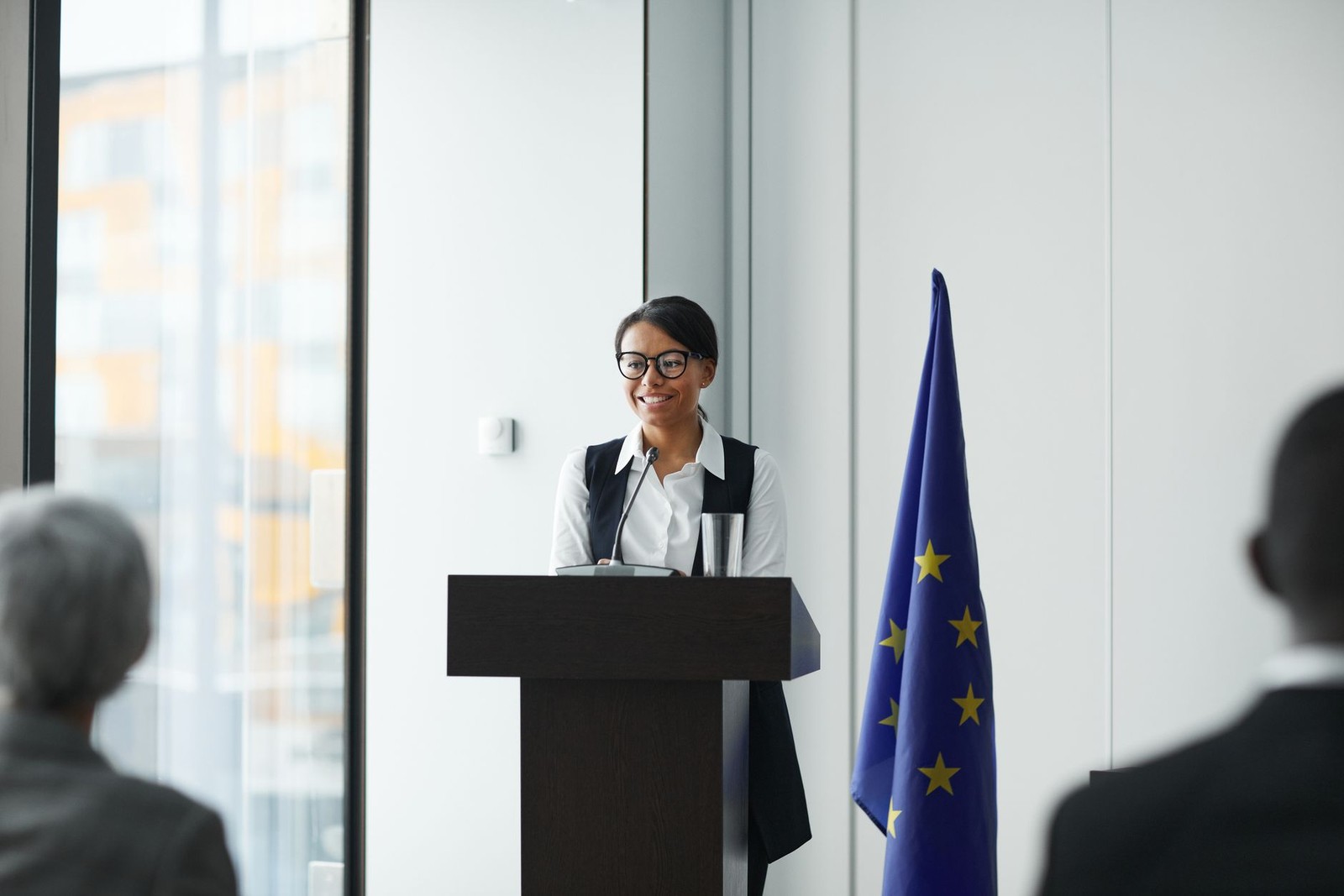 A woman in glasses smiles while speaking at a lectern beside a European Union flag, addressing seated attendees