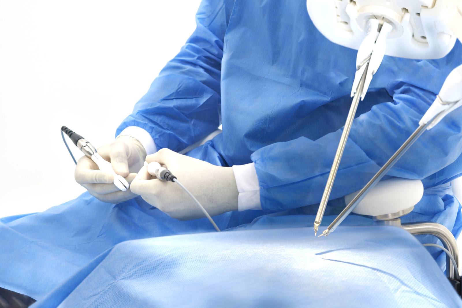 A gowned surgeon seated at the Symani Surgical System holds its two hand controllers while the robot's two slender wristed NanoWrist instruments reach down toward the draped surgical field