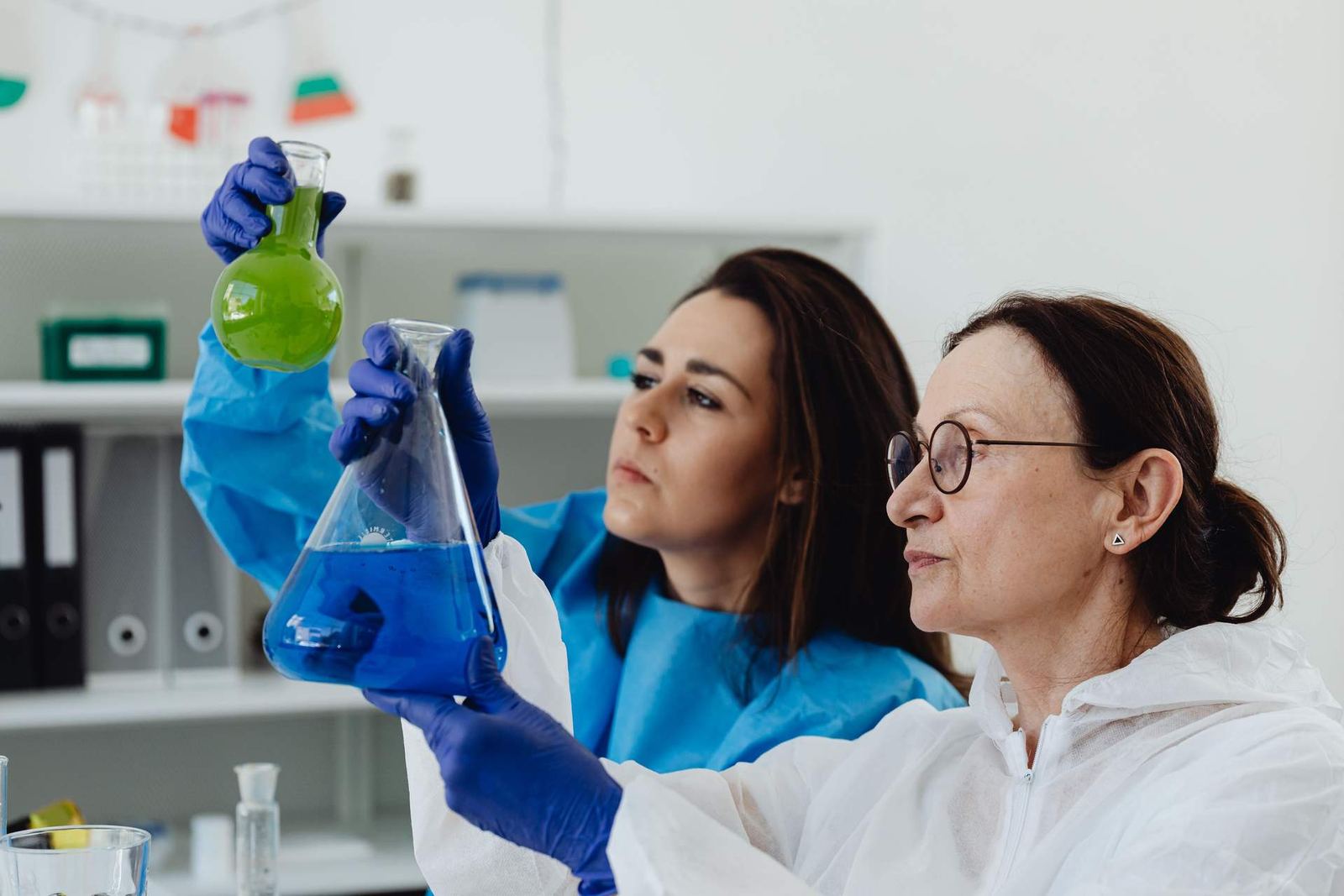 Two researchers in lab coats and nitrile gloves examine flasks of colored liquid during a laboratory review