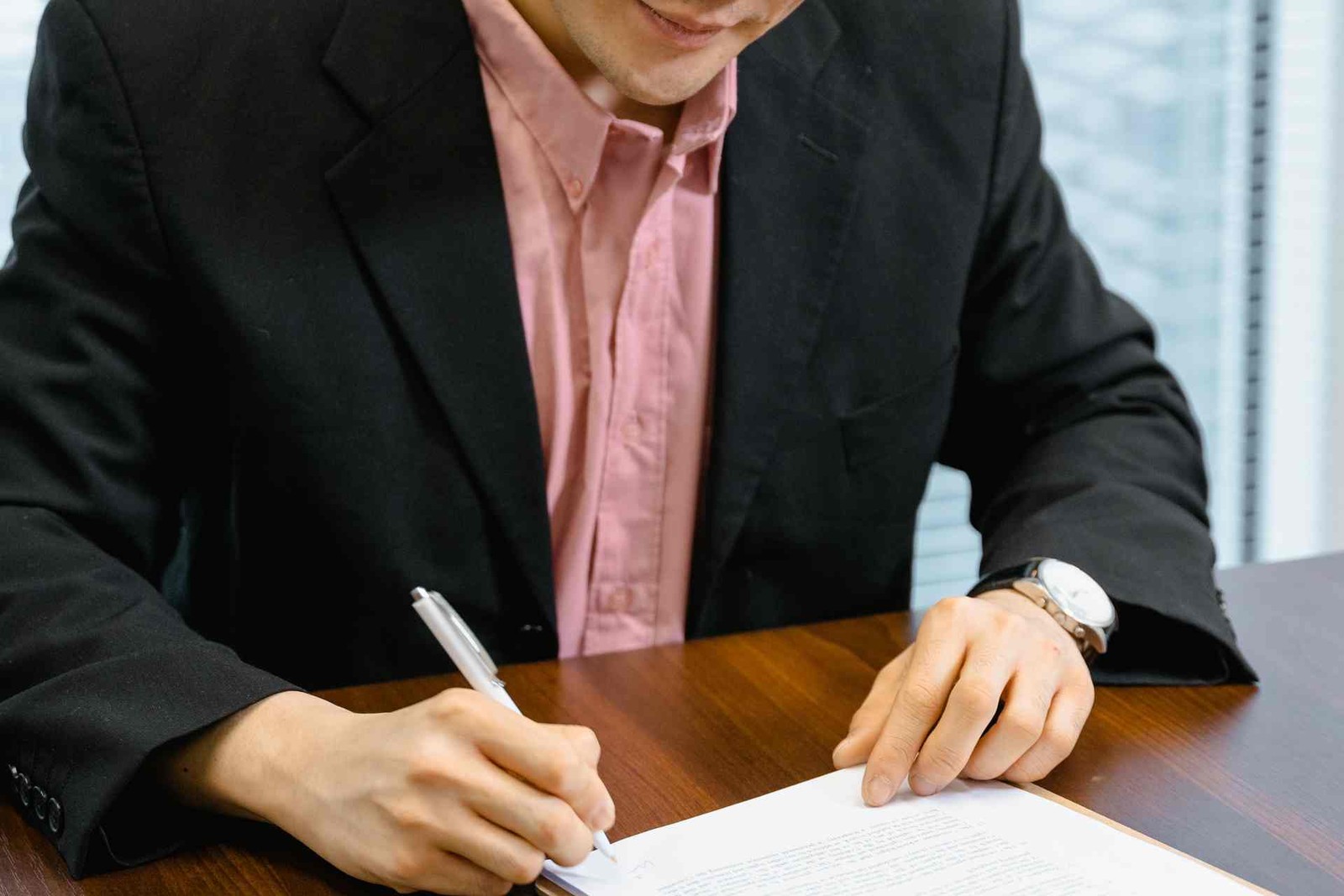 A close-up of a man in a suit signing a printed document with a pen at a wooden desk