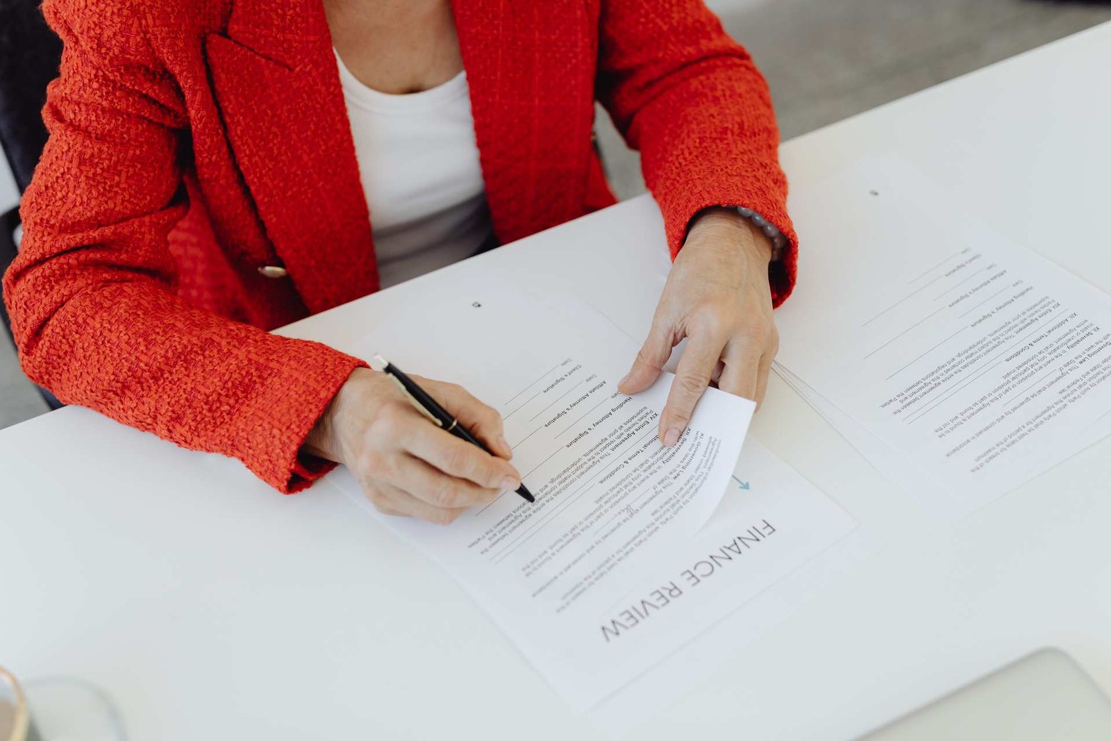 A person in a red blazer signs a paper form on a desk, pen in hand