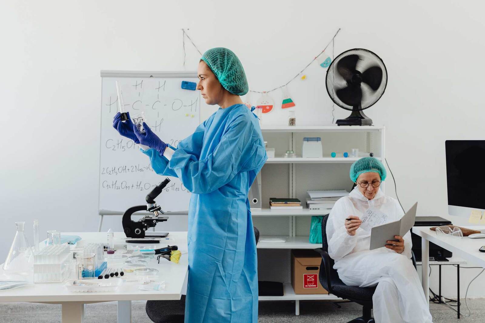 A scientist in a lab gown examines samples at a lab bench with a microscope and glassware