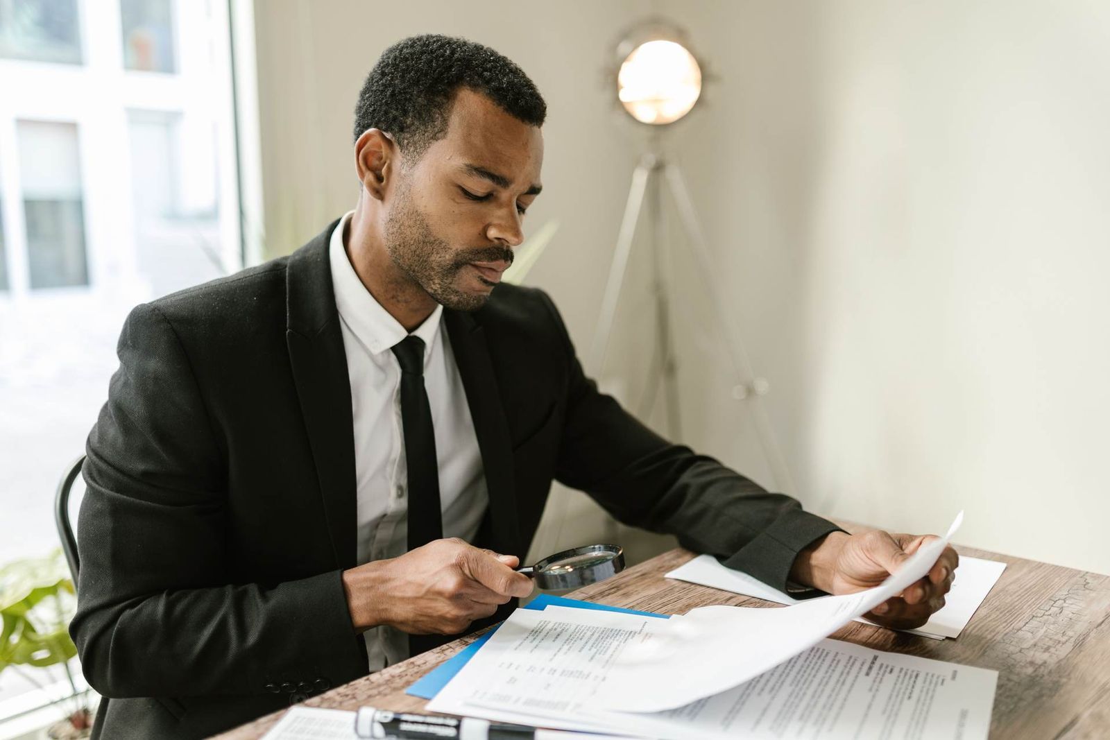 A man in a suit examines a document closely with a magnifying glass at a desk