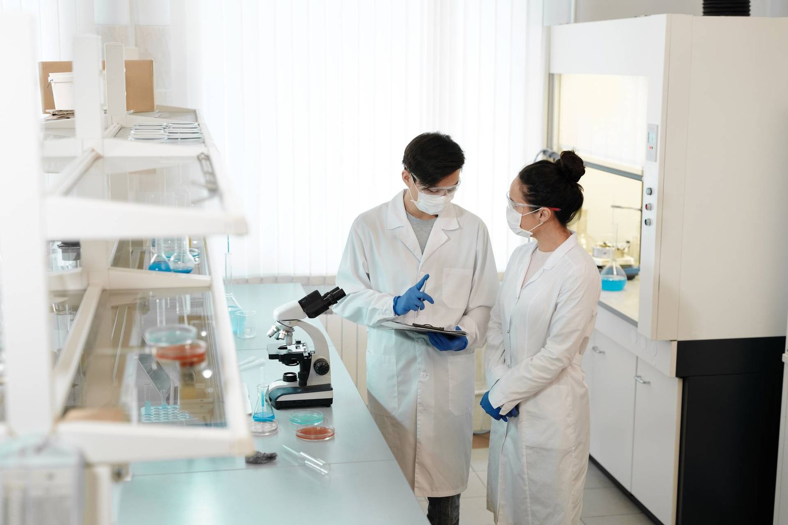 Two masked researchers in white lab coats confer over a clipboard in a laboratory, standing beside a microscope and glassware