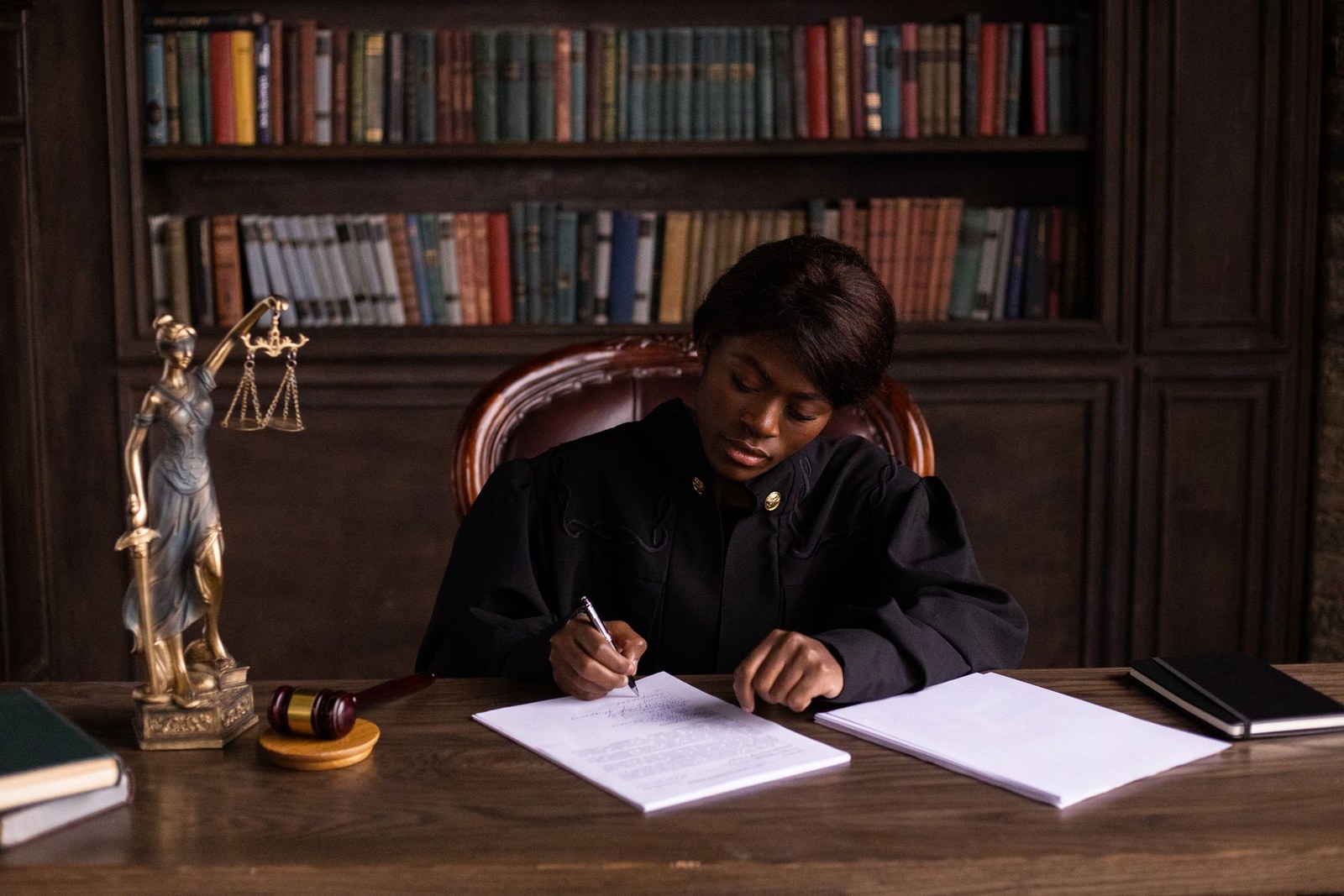 A judge in black robes signs a document at a wooden desk beside a statue of Lady Justice and a gavel