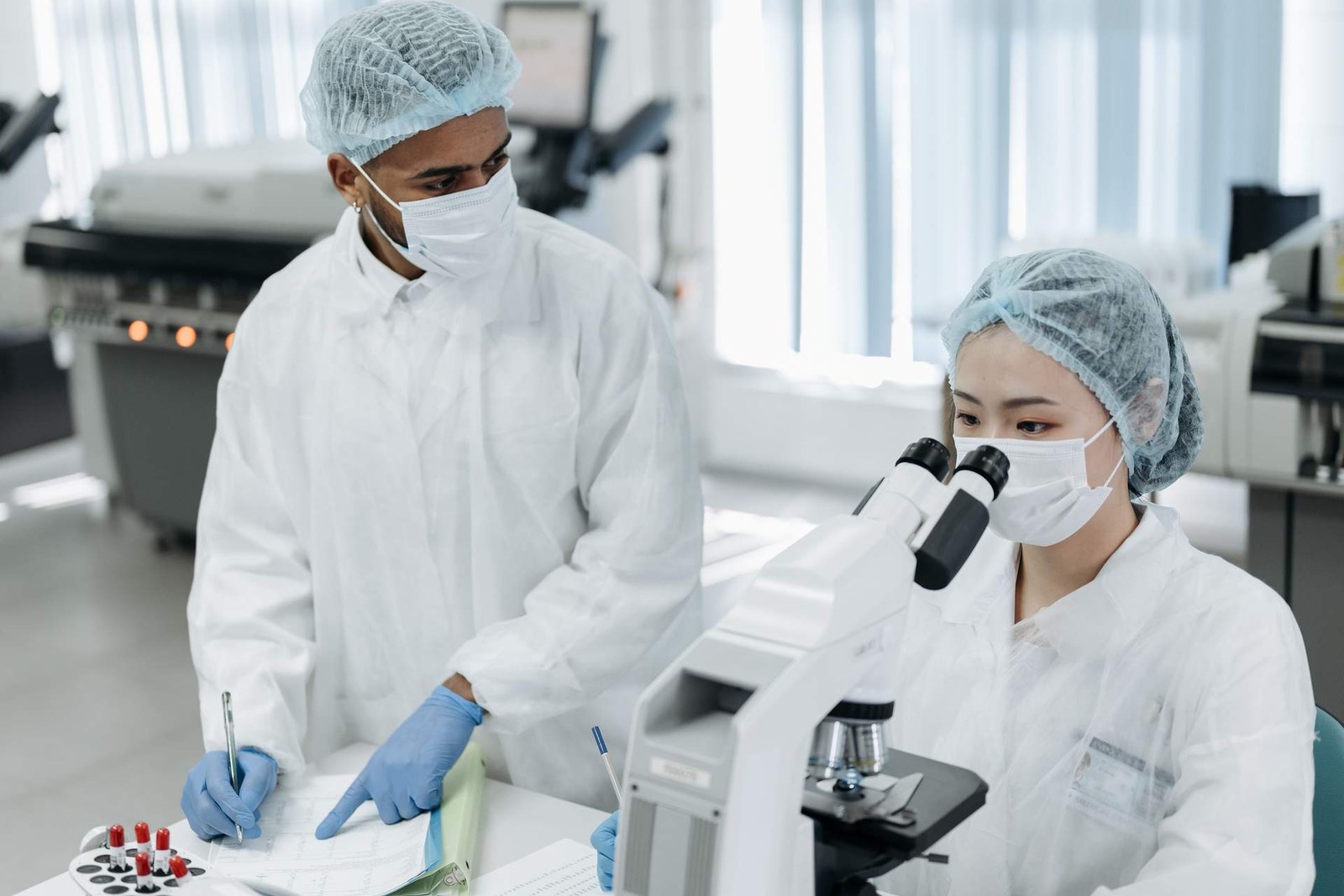 Two gowned, masked laboratory researchers in hairnets examining a sample under a microscope while taking notes
