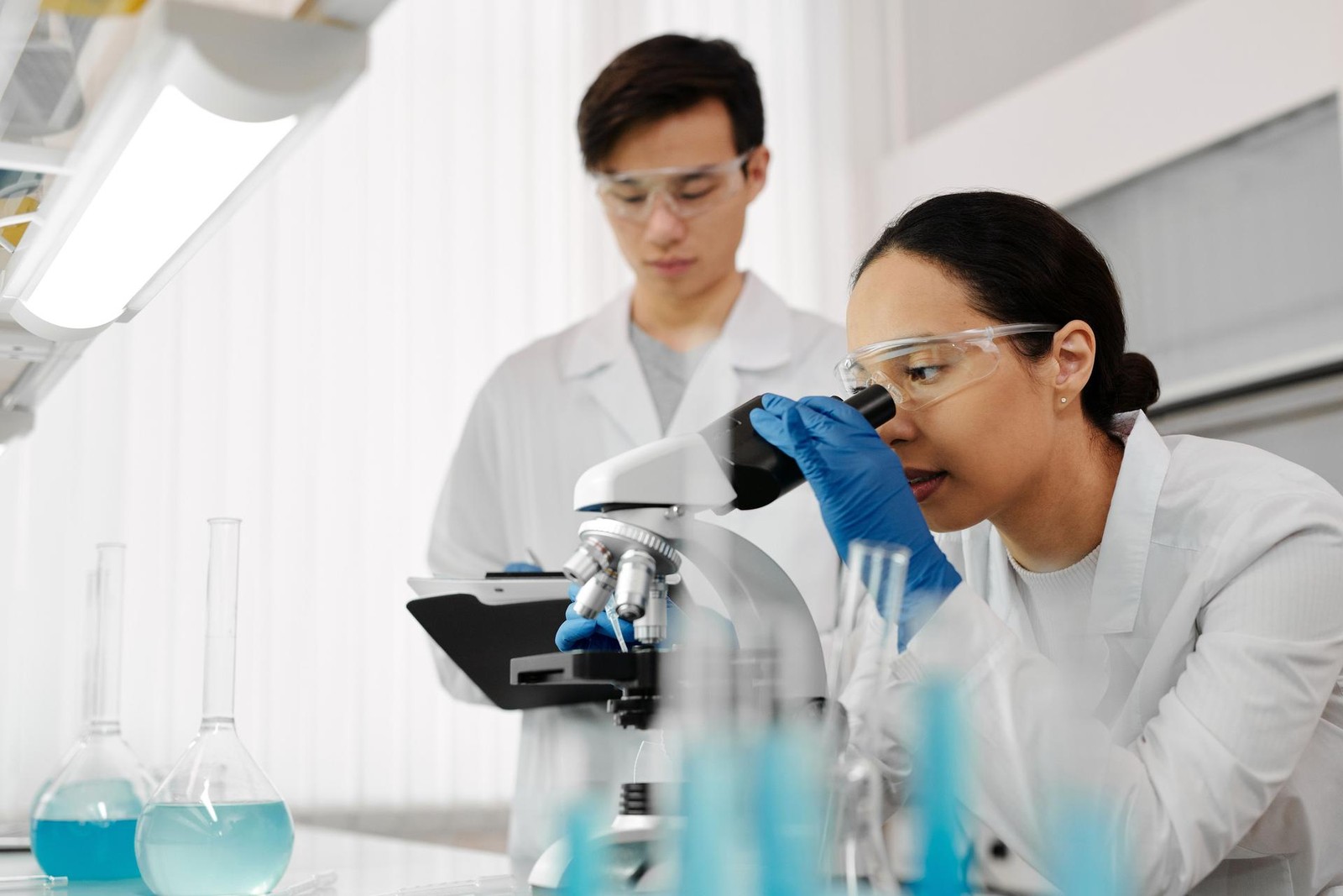 A researcher examines a specimen through a microscope while a colleague takes notes on a tablet in a bright laboratory