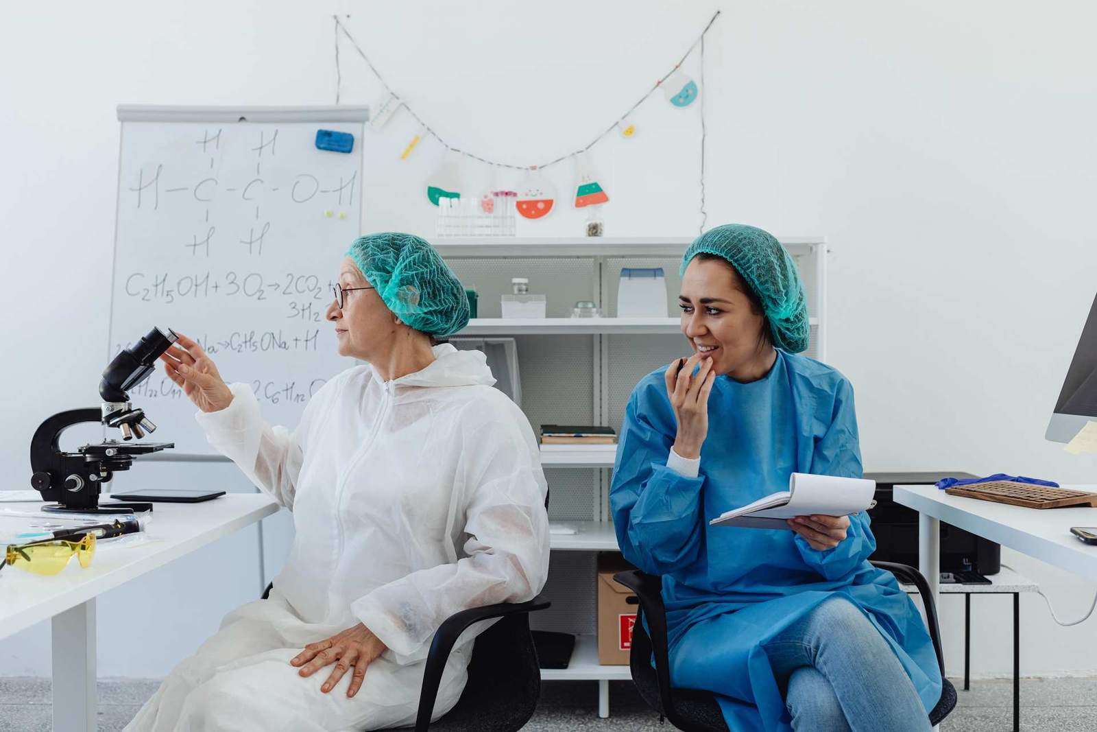 Two scientists in a laboratory, one in a white coat gesturing toward a microscope while the other, in blue scrubs, looks on and smiles