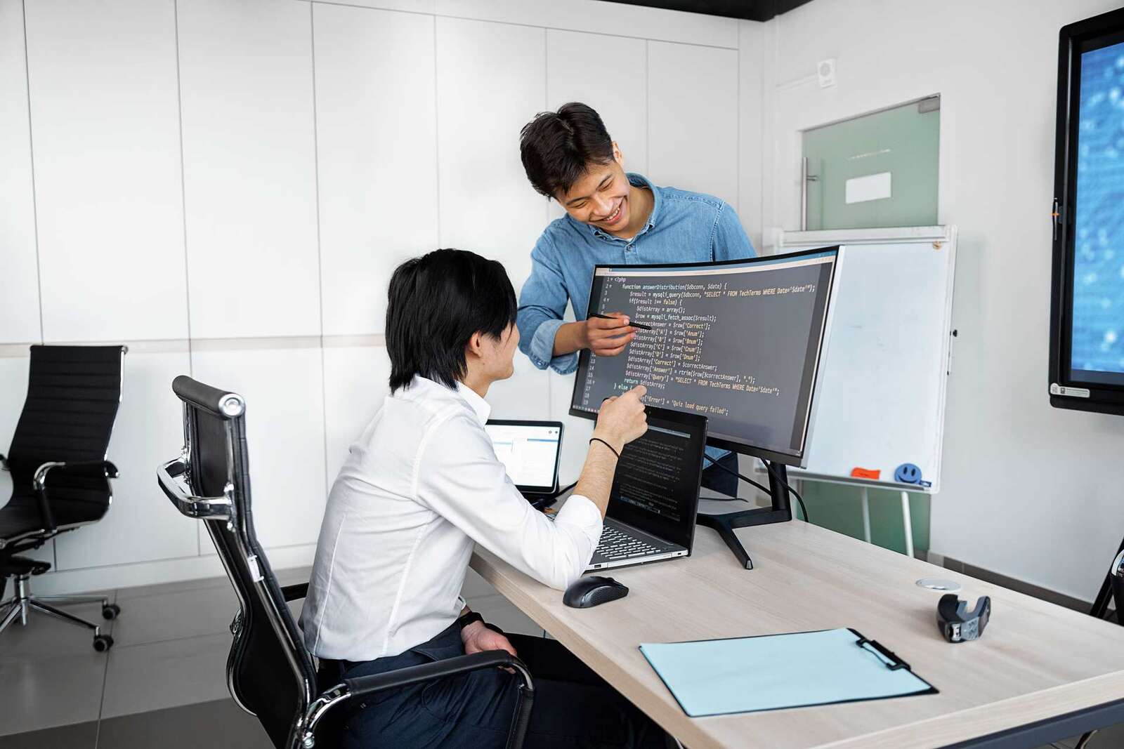 Two colleagues point at code and charts on a monitor in a bright office