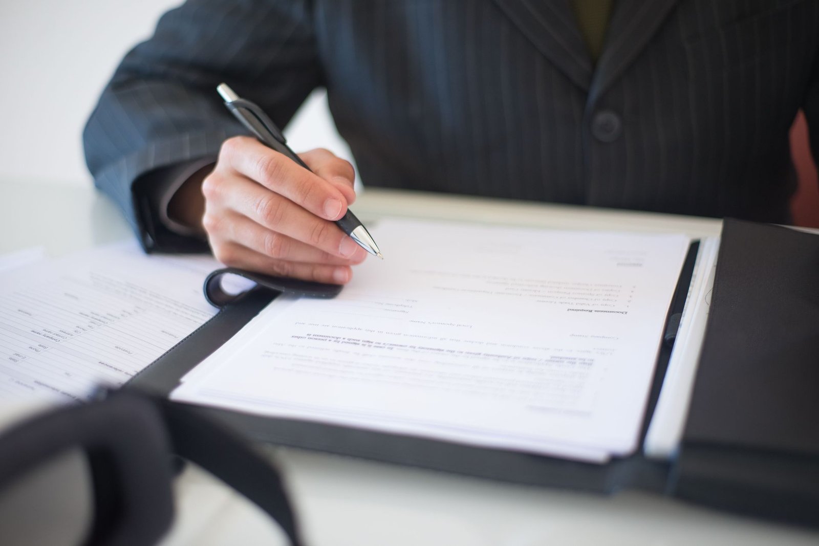 A close-up of a hand in a pinstripe suit signing a regulatory document on a clipboard