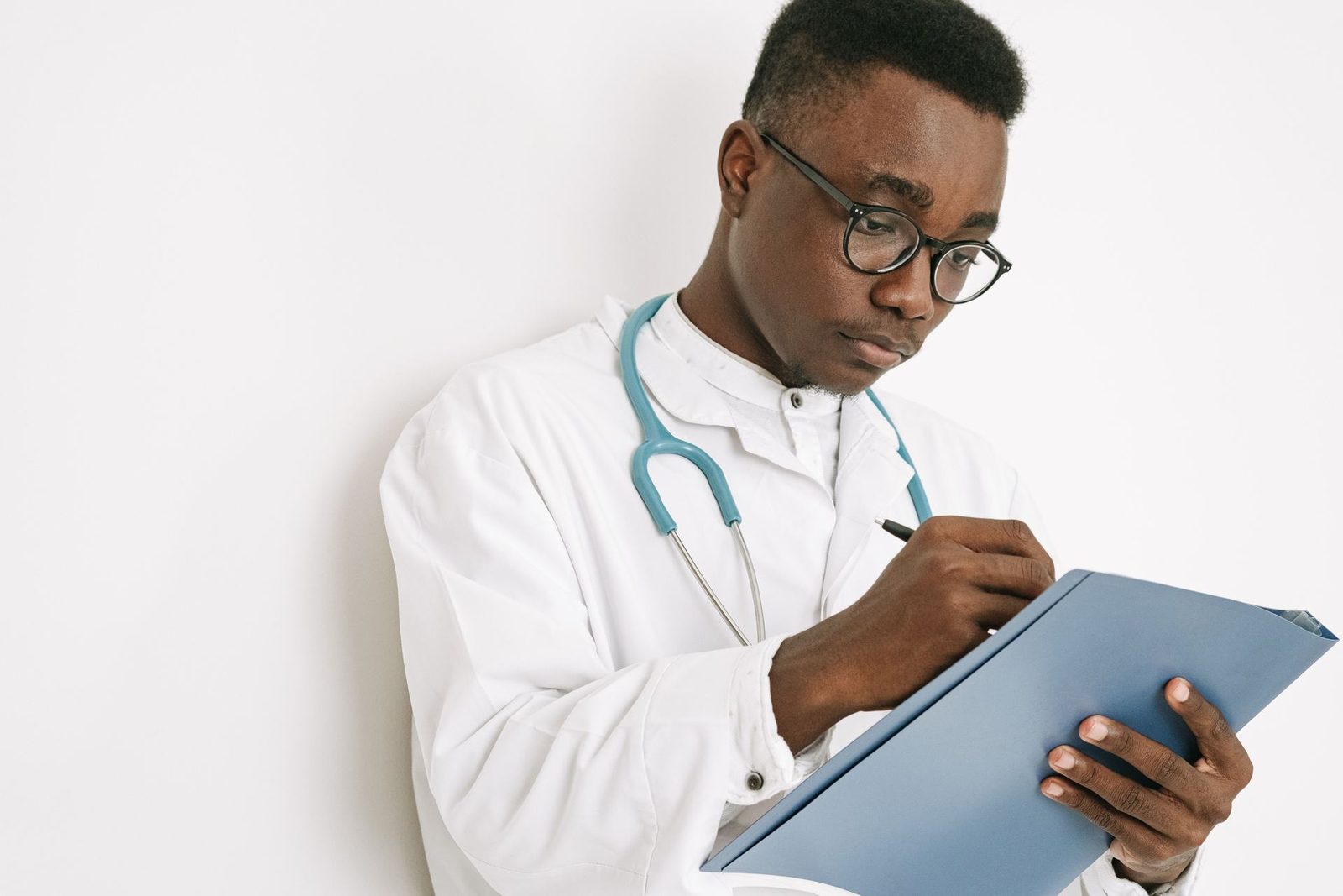 A physician in a white coat with a stethoscope writes notes on a folder against a plain studio background