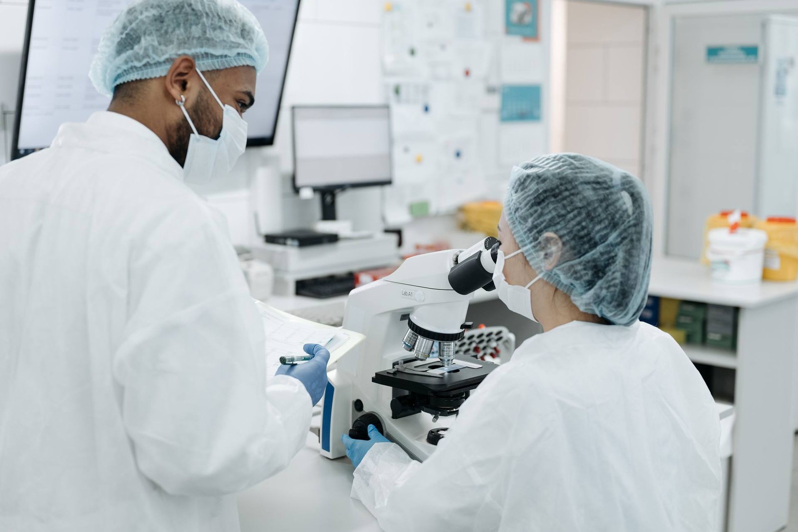 Two gowned technicians in hairnets and masks conferring over a microscope and lab notes in a cleanroom