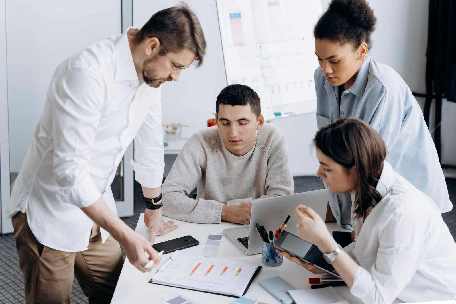 Four colleagues review a bar chart and data on a laptop around a white desk