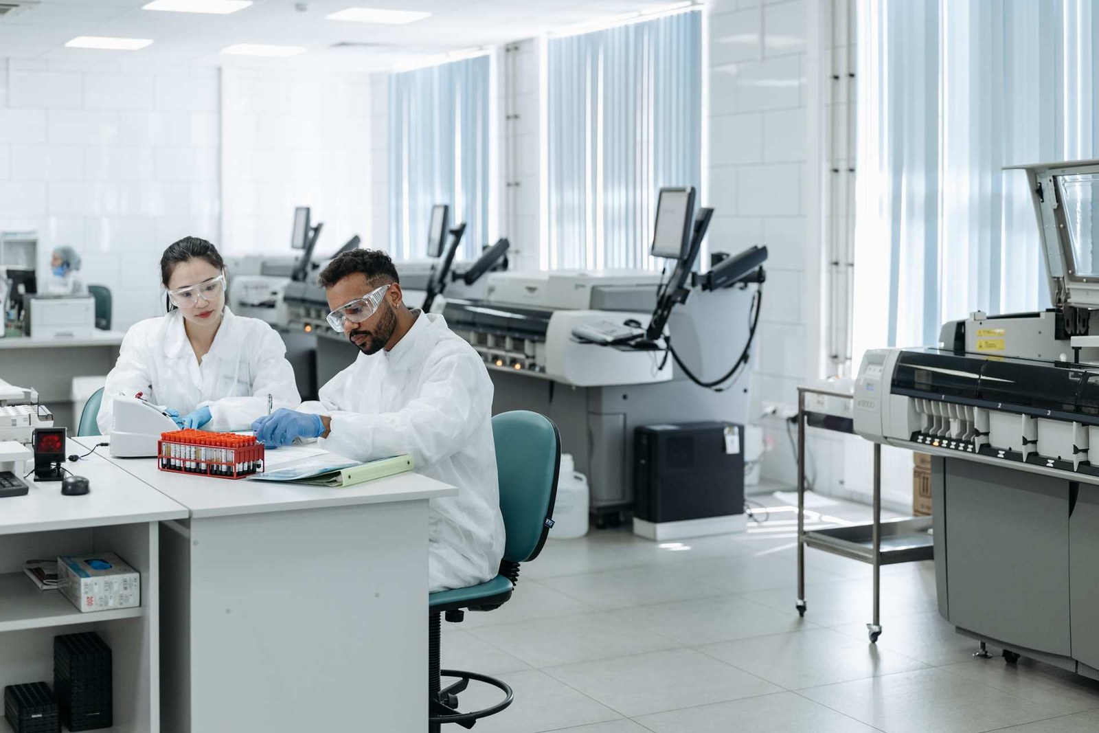 Two lab technicians in a clinical laboratory review blood sample vials and analyzer readouts