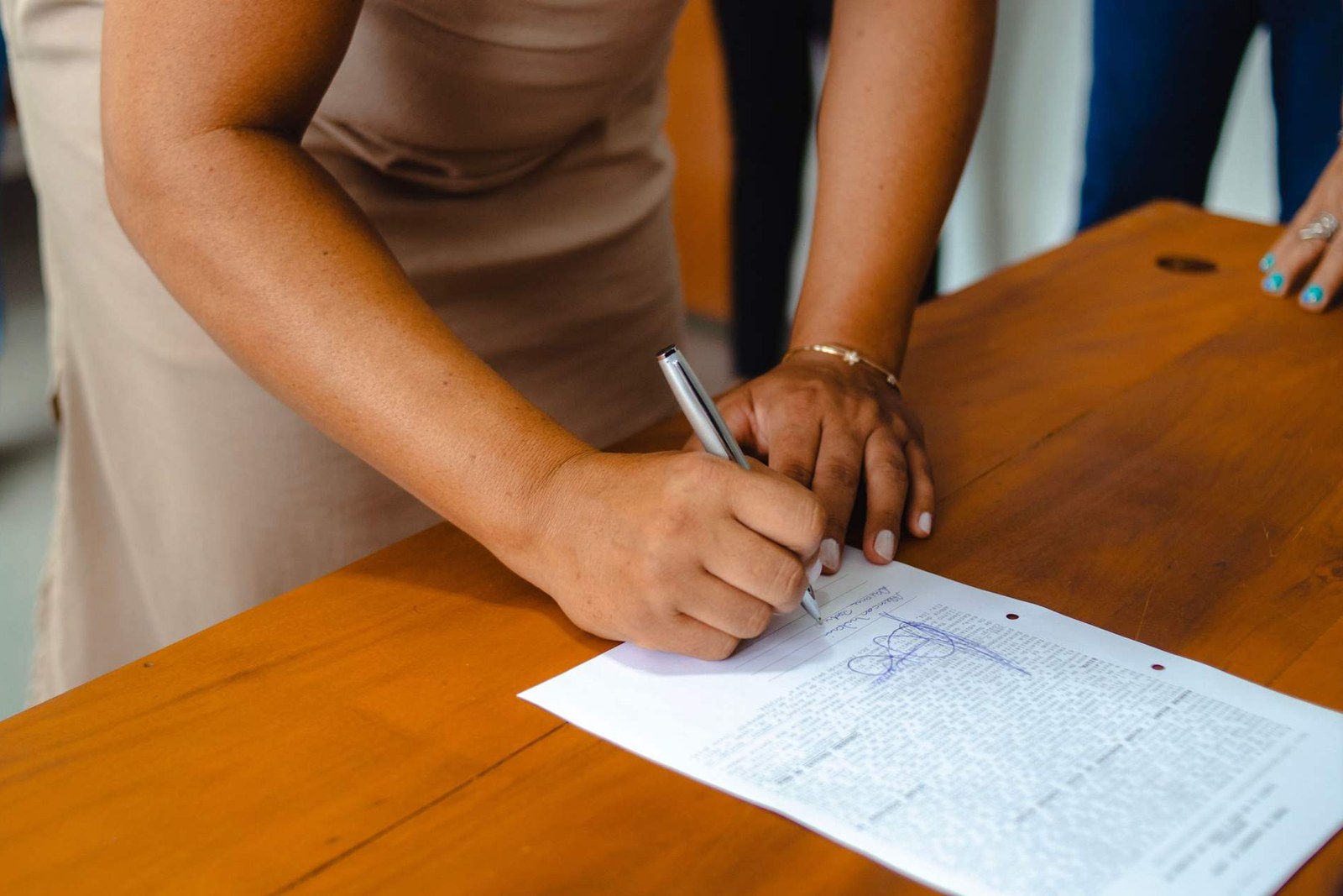 A person signs a printed document by hand with a pen while a colleague stands nearby at a wooden table