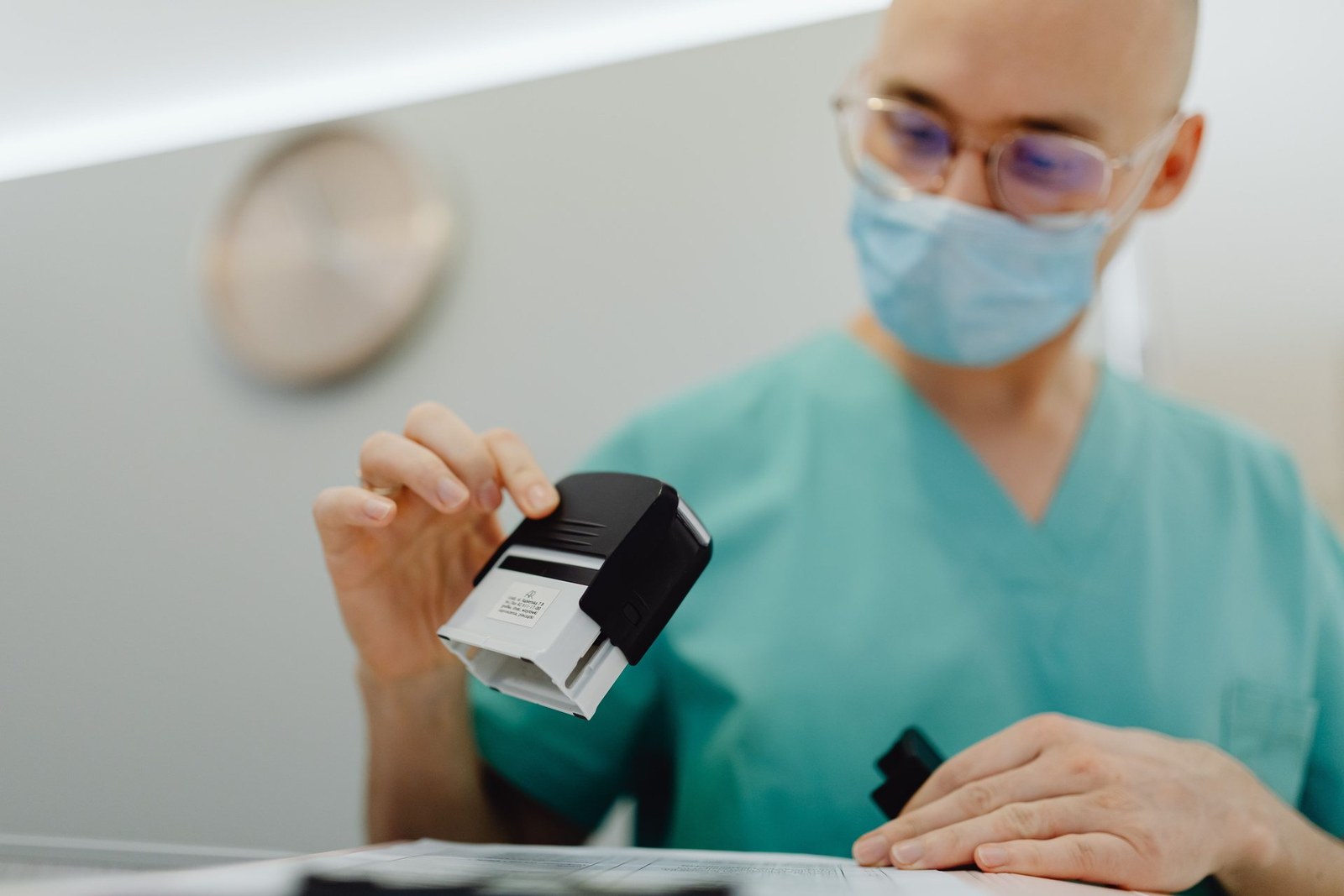 A masked clinician in scrubs examines a handheld stamping device over paperwork on an exam-room counter