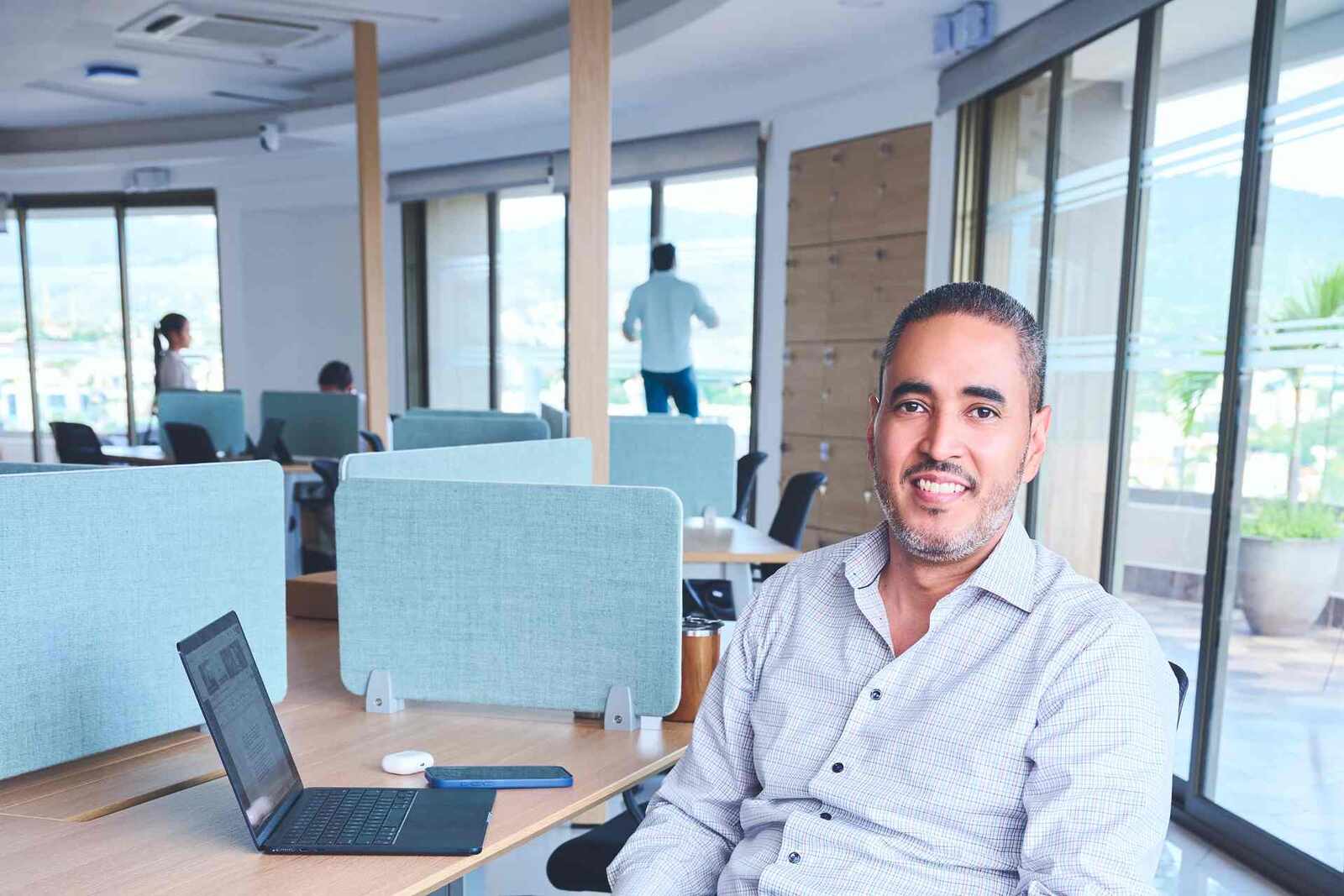 A solo executive in a checked shirt smiling at his desk in a bright, glass-walled open office