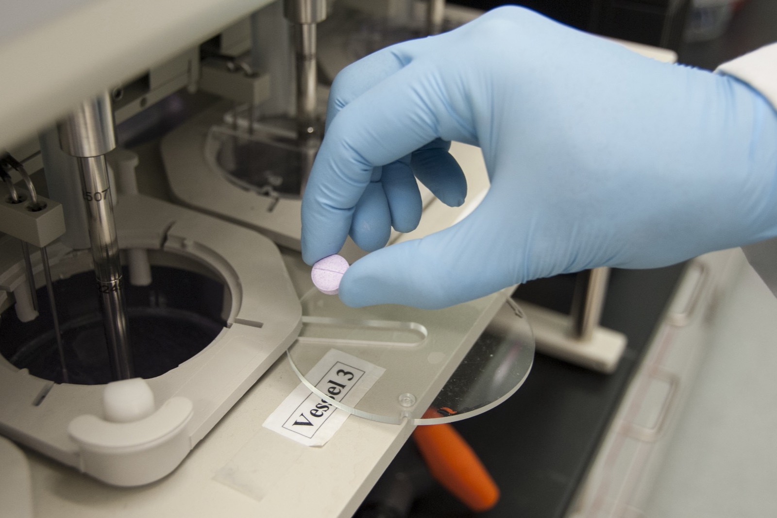 A gloved hand holding a tablet over dissolution-testing equipment in an FDA laboratory