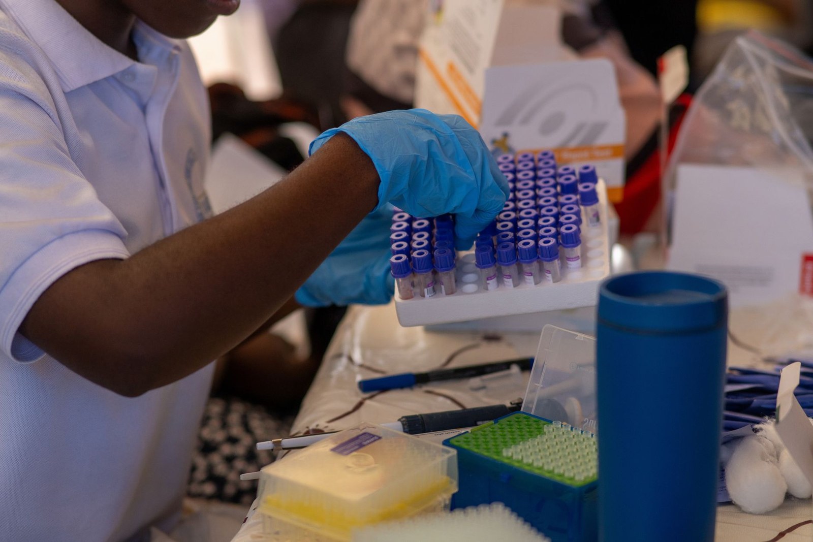A gloved hand lifts a rack of purple-capped sample collection tubes at a testing table, with a rapid-test box in the background