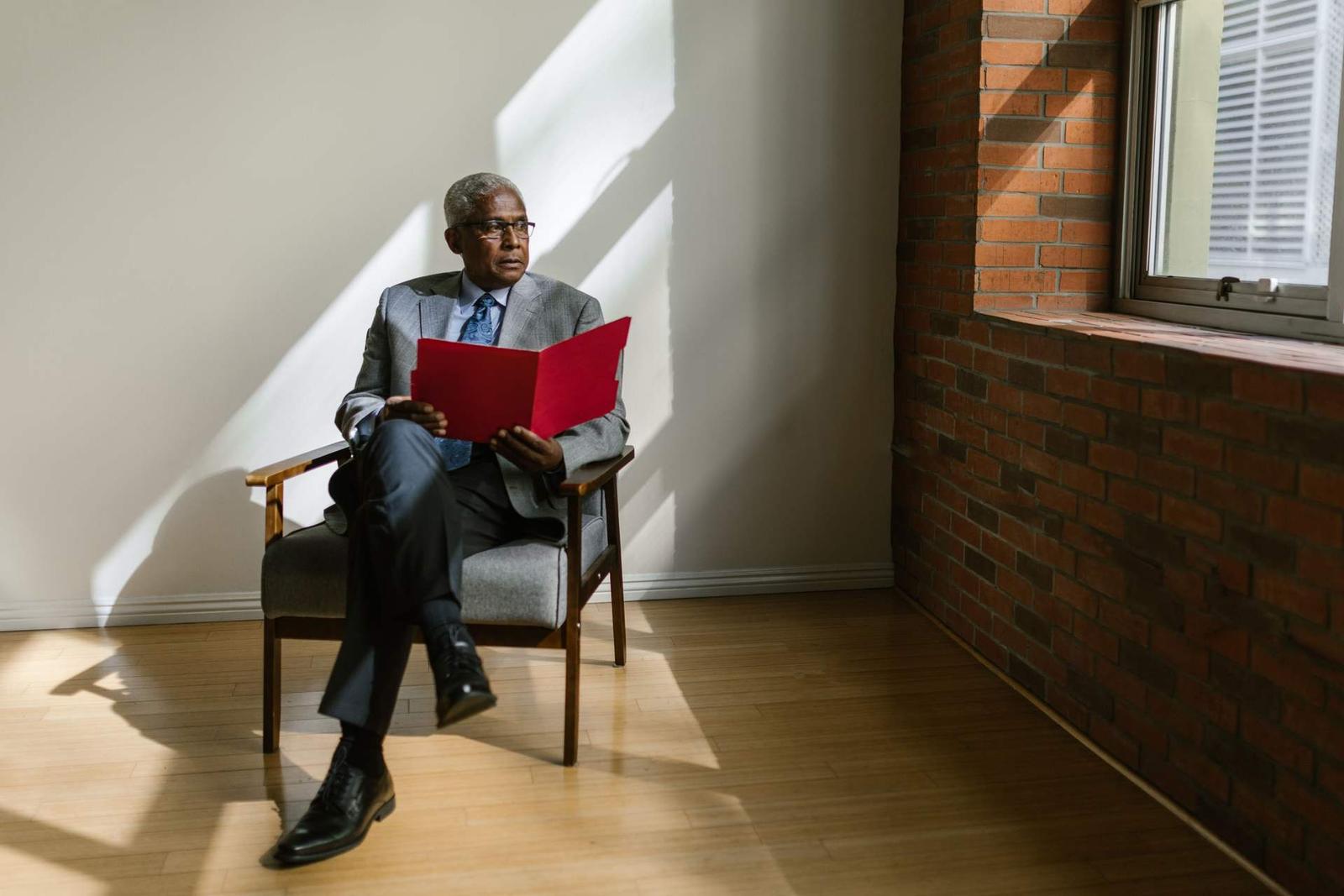 A man in a grey suit sits in a sunlit chair reading from an open red folder, near a window and a brick wall
